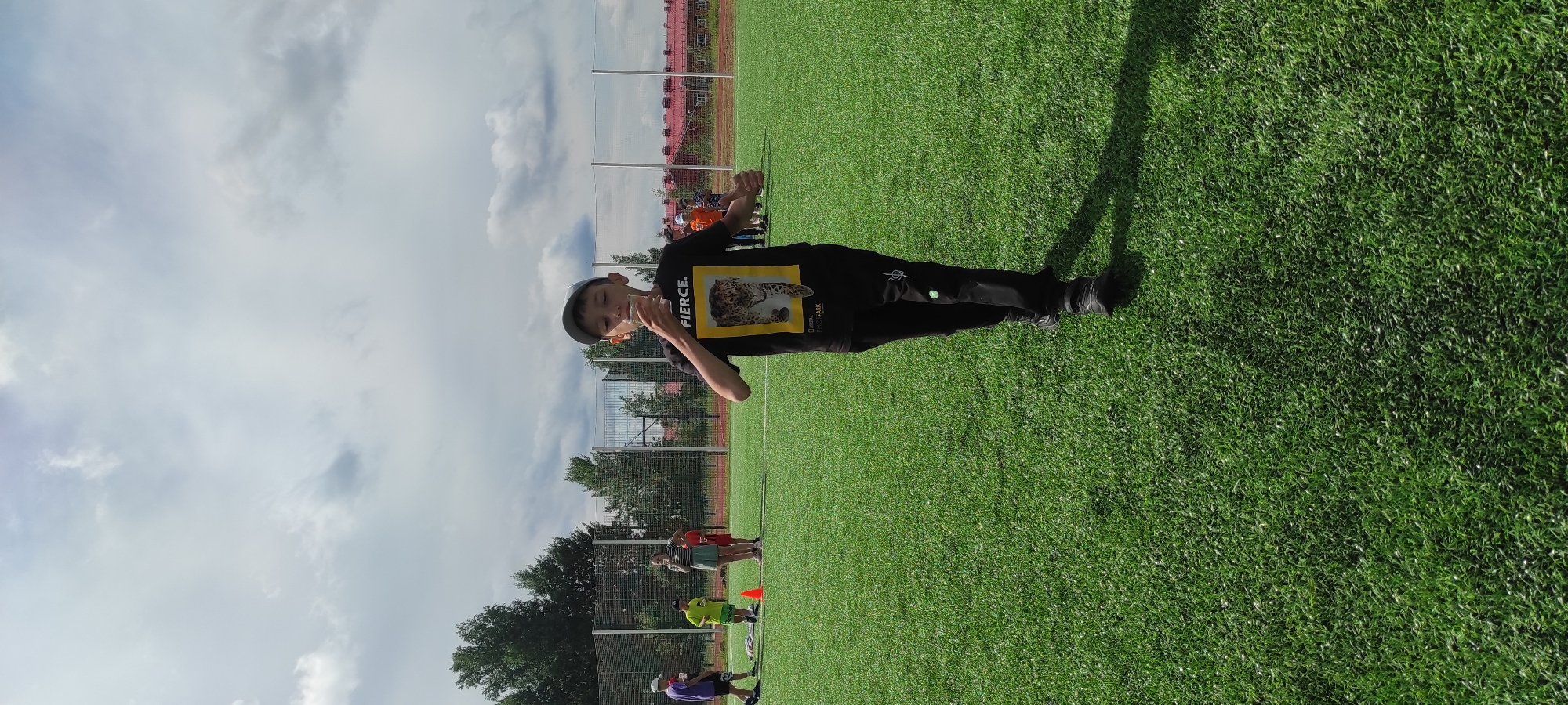 A young boy in a black t-shirt and black pants is walking across a green turf field, holding a clear cup to his lips. He is wearing a gray baseball cap. The t-shirt has a graphic of a leopard and the word "FIERCE" on it. In the background, several other children and adults are visible, some standing and some walking. There are soccer cones and a net in the distance, suggesting this is a sports field or park. The sky is partly cloudy. The location is identified as Noviy, Russia.