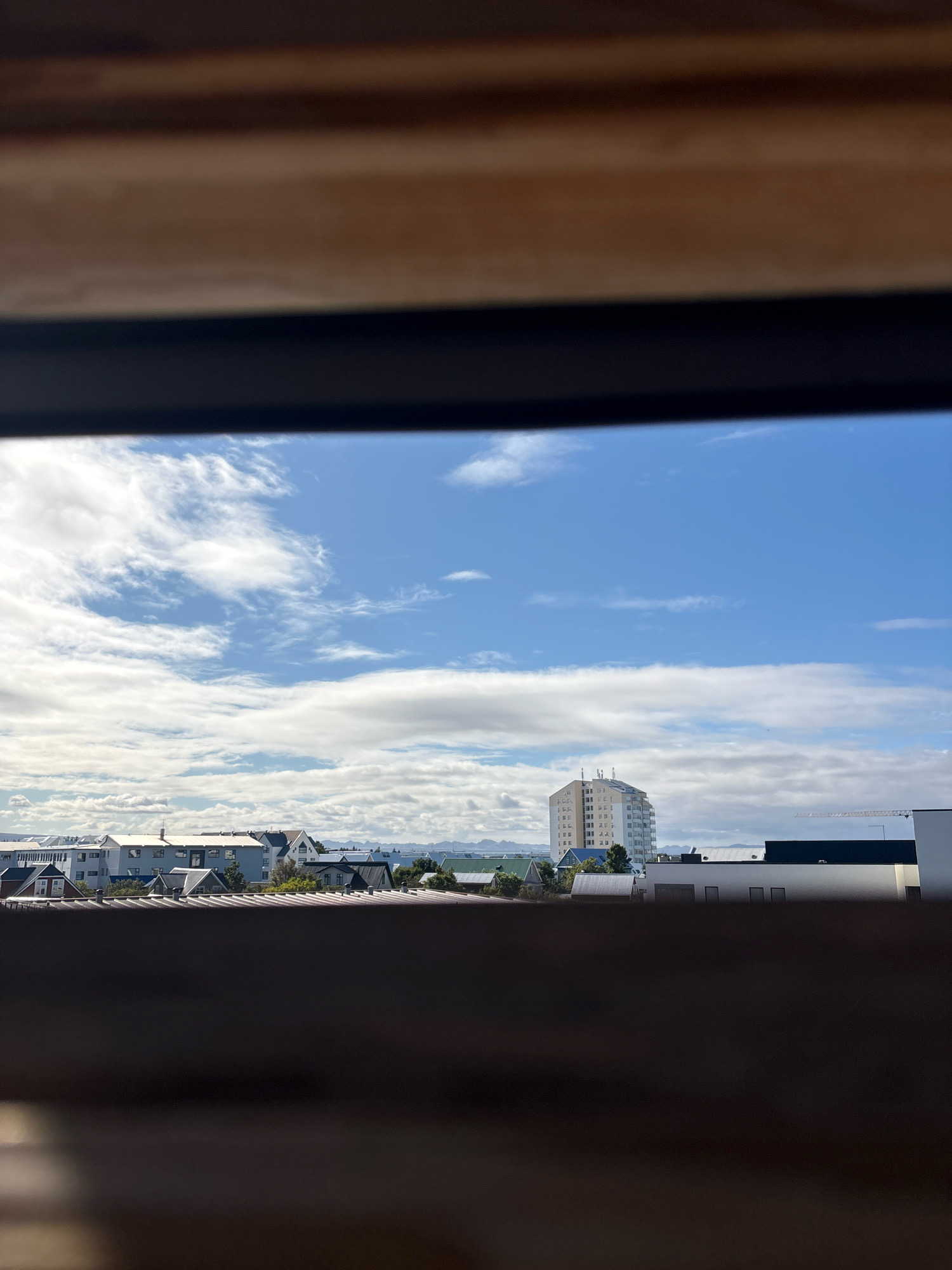 This is a daytime, outdoor, eye-level shot of a cityscape in Reykjavik, Iceland, viewed through a wooden frame. The foreground is dominated by a blurred, dark wooden structure, likely a window frame or railing, obscuring the bottom third of the image. Beyond this, the middle ground displays a collection of buildings of varying heights and architectural styles. In the background, a tall, modern, light-colored apartment building stands out, with a light blue sky above, dotted with white, wispy clouds. Further in the distance, a range of mountains can be seen, their peaks lightly dusted with snow. The scene is bathed in natural daylight, indicating it was captured during daytime hours.
