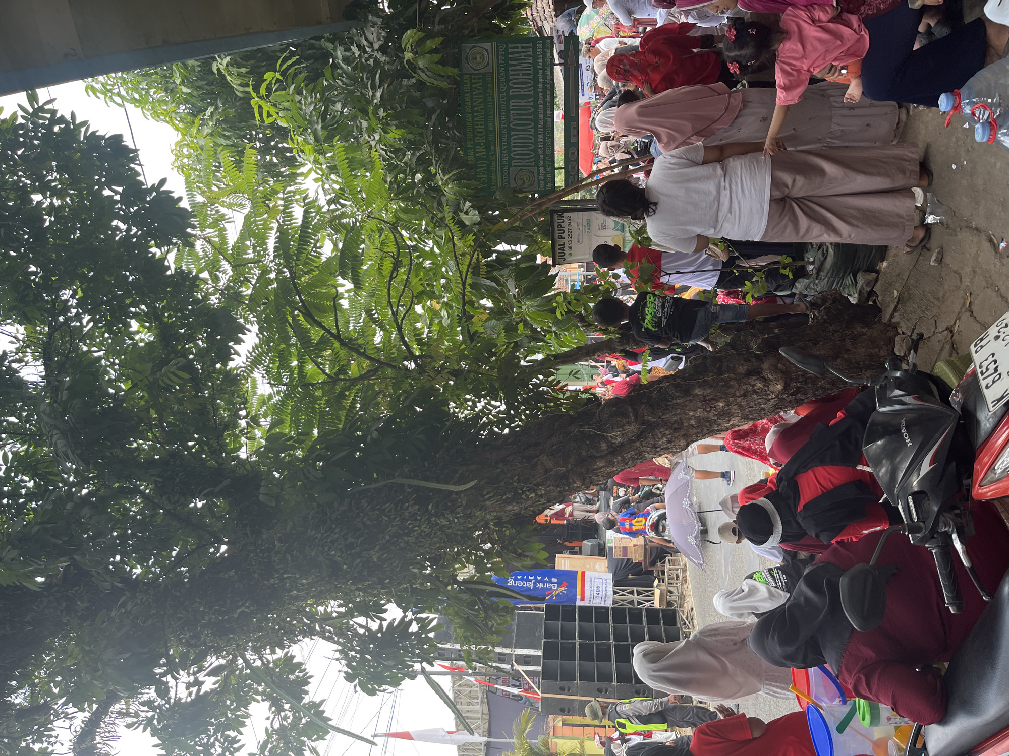 The image depicts an outdoor scene in Glagah Kulon, Indonesia, likely during an event. A large tree with dense foliage dominates the left side of the frame, partially obscuring the view of a stage area with large speakers on the left. Several people, many wearing hijabs and traditional Indonesian attire, are gathered. Some are seated on the ground, while others stand. In the foreground, a motorcycle is parked. A signpost in the center-right reads "JUAL PUPUK" (fertilizer for sale) and another sign indicates "ROUDLOTUR ROHMAH" with an address in Glagah Kulon. There are also Indonesian flags visible. The overall atmosphere suggests a community gathering or market.