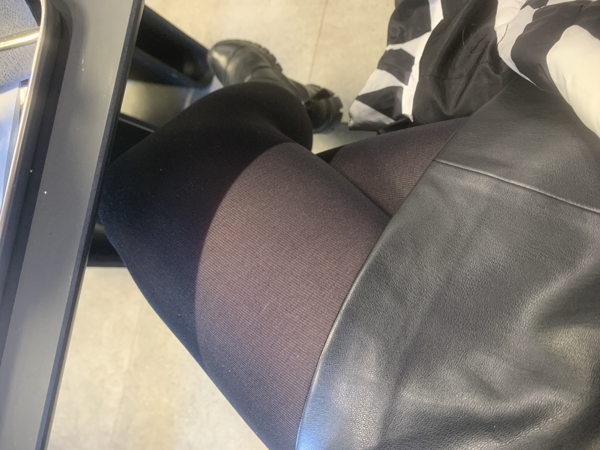 The image shows a person sitting on a chair, wearing black boots and leggings with what appears to be a black leather skirt or jacket. A portion of a black and white patterned object is also visible near the person's right side. The environment consists of a light-colored floor and a portion of the chair. No information suggests that the location is Izhevsk, Russia.