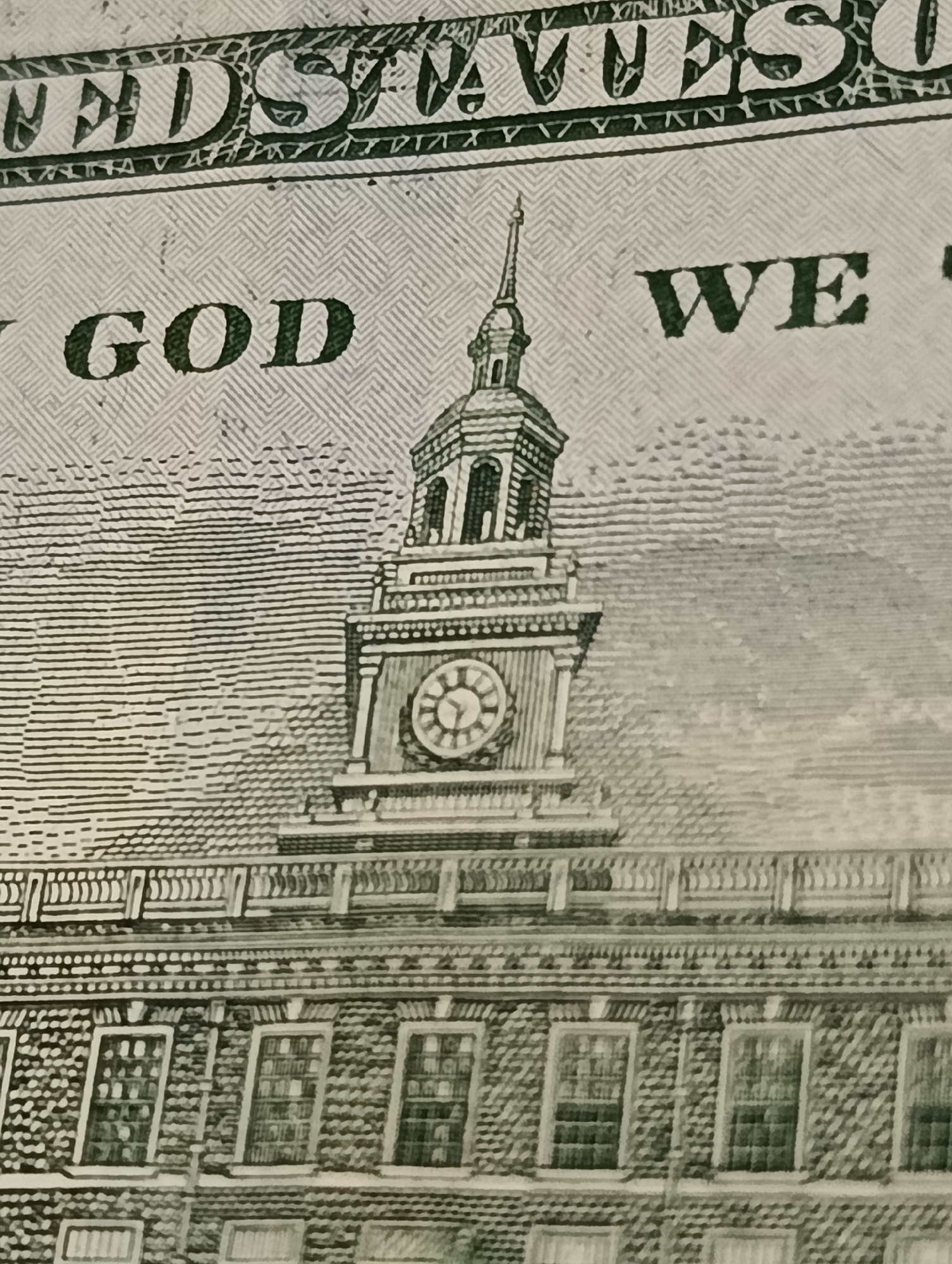 This is a close-up, macro shot of the back of a United States hundred-dollar bill. The image features the Independence Hall building in Philadelphia, Pennsylvania. Prominently displayed are the words "GOD" and "WE" in large, dark green, serif font, part of the inscription "IN GOD WE TRUST". The architectural details of the building are visible, including windows, a clock tower with a steeple, and decorative elements. The background consists of the intricate, textured security printing characteristic of U.S. currency. There are no people or actions depicted in the image.