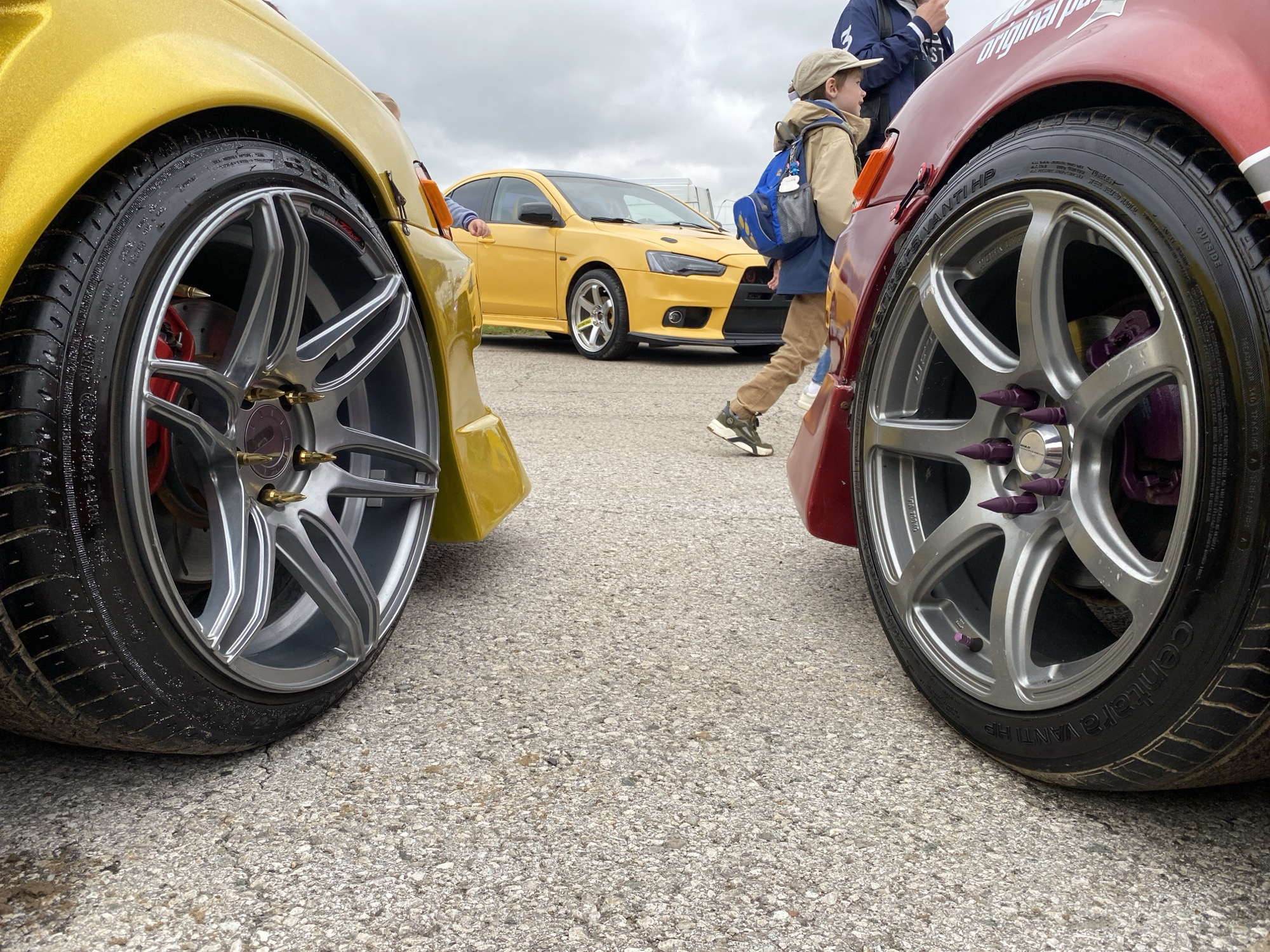 The image shows a low-angle, close-up view of the front wheels of two cars, a yellow and a red one, parked on a grey asphalt surface. The yellow car is on the left, and the red car is on the right. Both cars have custom rims with brightly colored lug nuts. In the background, a yellow Mitsubishi Lancer Evolution is parked. A young boy wearing a cap and a backpack is walking past the cars. The scene appears to be outdoors, possibly at a car show or event. The sky is overcast, suggesting a cloudy day. The image does not contain any elements that would specifically place it in the city of Perm, Russia.