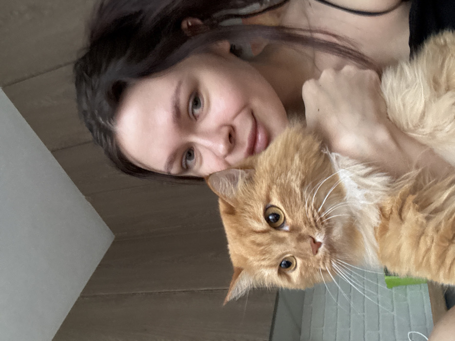 A woman with brown hair and fair skin smiles into the camera while holding an orange cat. The cat has wide eyes and long, white whiskers. The background includes a wooden wall and a white wall. The woman's dark hair is pulled back, and she is wearing a black tank top. The photo is taken at an angle, with the woman and cat tilted slightly.