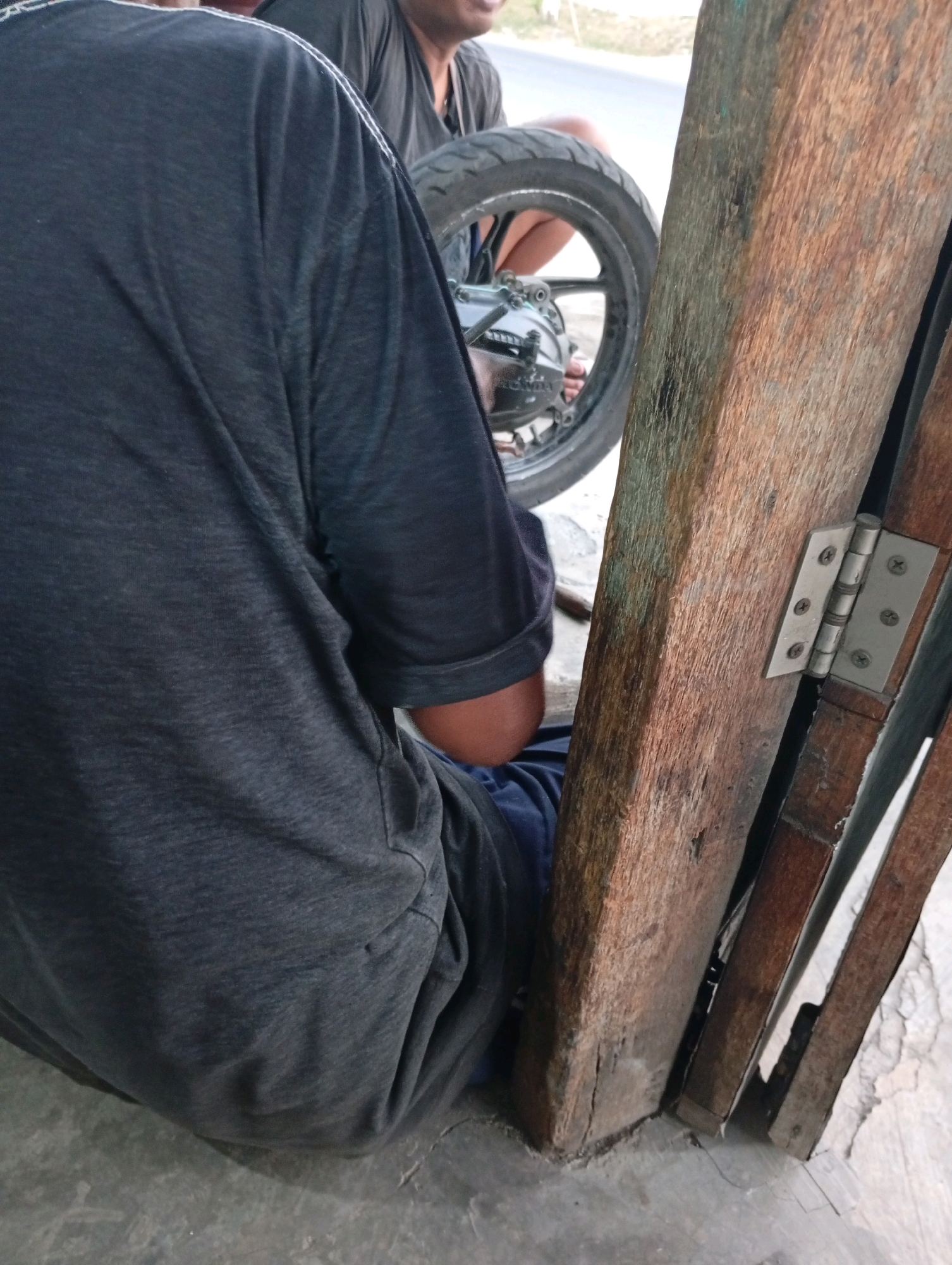 The image shows a person sitting next to a motorcycle tire, with their back to the camera. The person is wearing a dark gray t-shirt and dark blue pants. They are leaning against a wooden structure that appears to be a part of a building or a gate. A metal hinge is visible on the wooden structure. The ground is made of concrete. The background shows a road and some vegetation. The image is taken from a low angle. The location is identified as Banyu Ajuh, Indonesia.