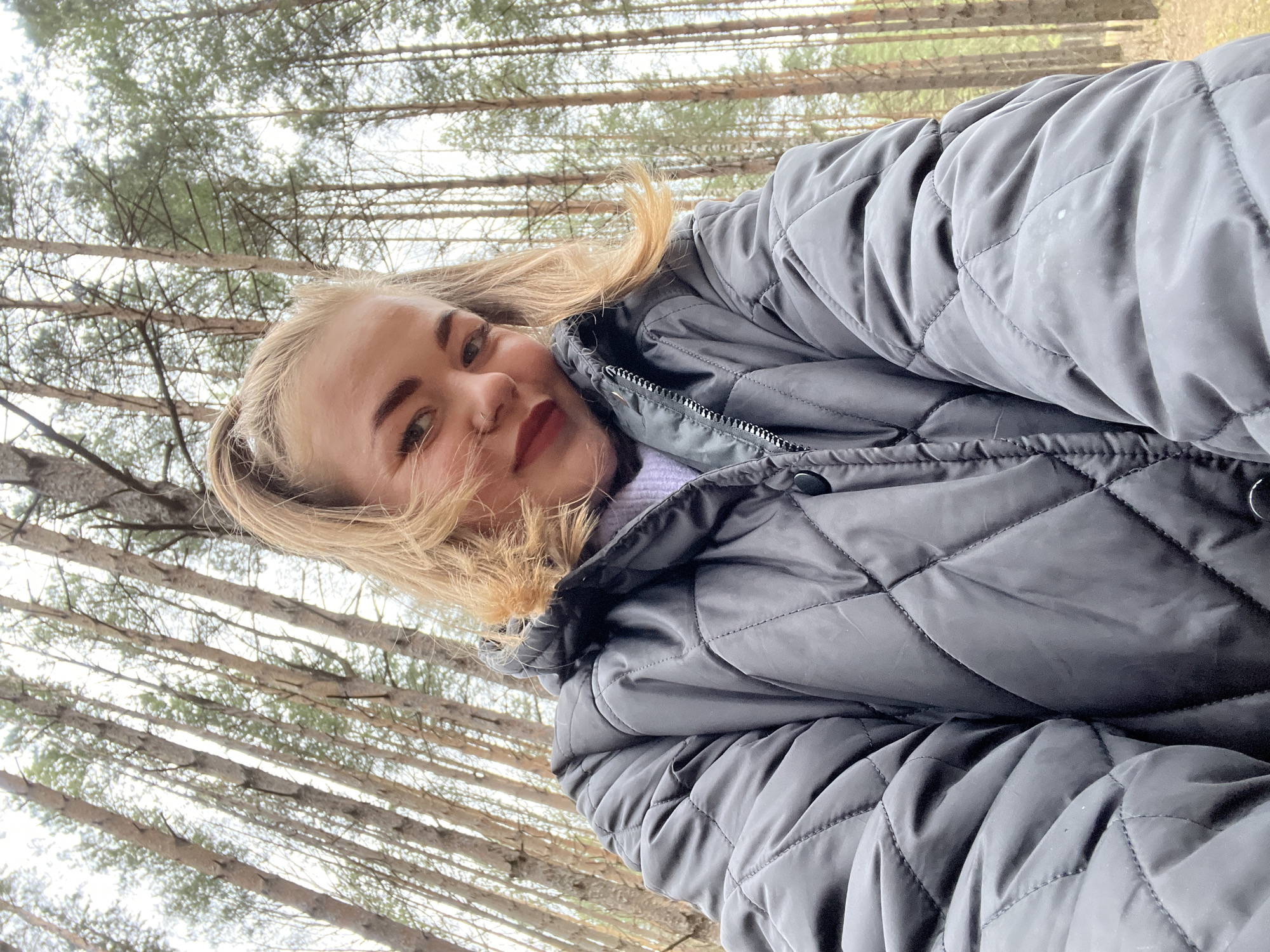 A selfie captures a young woman outdoors in a forest in Chaikovsky, Russia. The woman, with blonde hair, light skin, and dark eyebrows, is seen from the chest up, smiling gently at the camera. Her hair is slightly windswept, framing her face. She is wearing a dark grey, quilted, puffy jacket with a zipper, layered over a light purple top, suggesting cool or cold weather.
The background consists of a dense coniferous forest with numerous tall, slender tree trunks reaching upwards. The foliage on the trees appears sparse, indicating either late autumn or winter. The sky above is bright and overcast, creating soft, diffused lighting across the scene. No specific activity beyond taking the selfie is depicted. There is no visible text in the image. The overall mood conveyed by the woman's expression is relaxed and pleasant.
