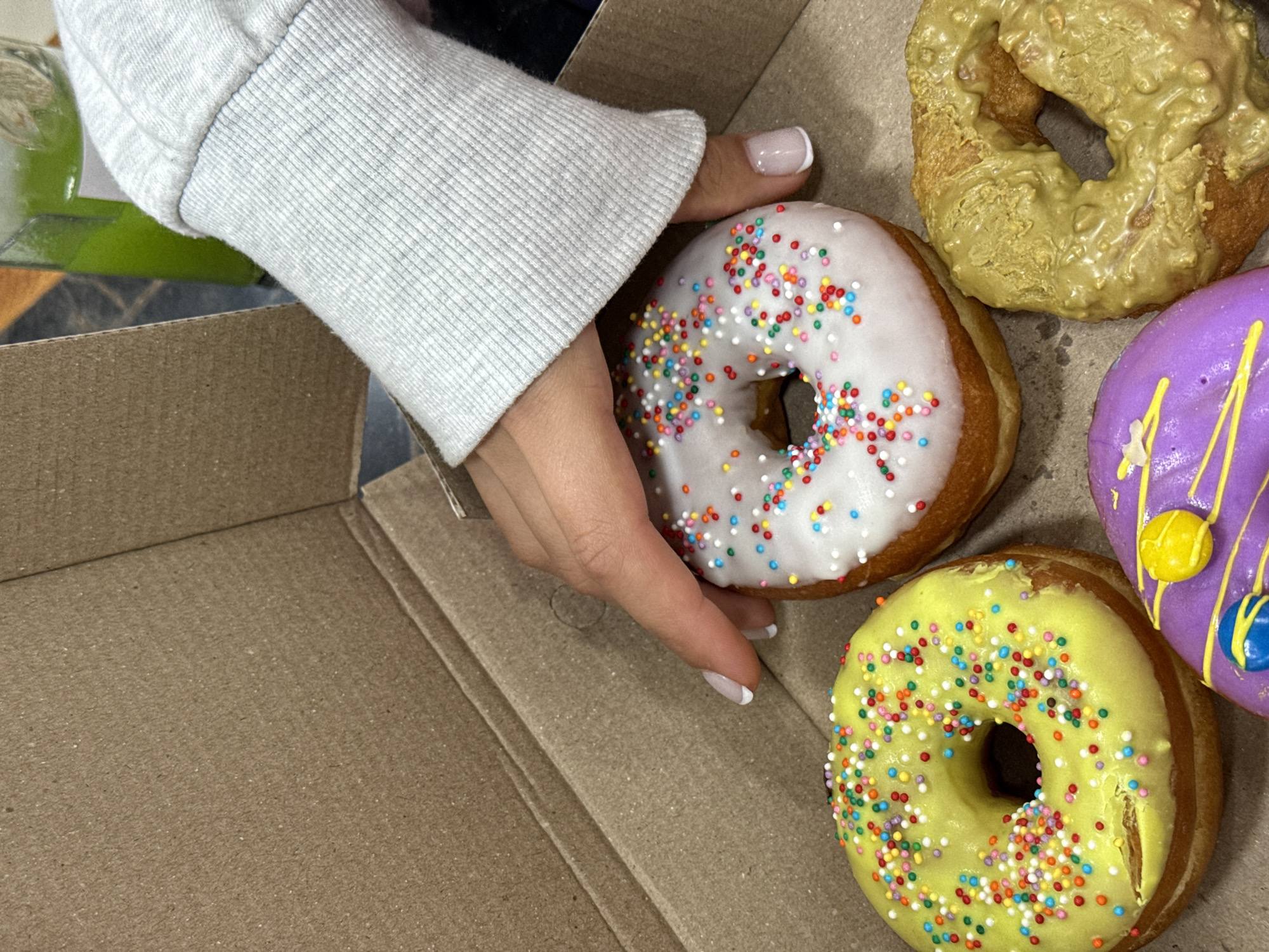 A close-up shot depicts a partly visible person holding a donut above a cardboard box filled with four colorful donuts. The person has fair skin, manicured nails with white tips, and is wearing a gray sweater. The donut they are holding has a white glaze and is covered in colorful sprinkles. Inside the box, there is a donut with a greenish-yellow glaze and another with a yellow glaze, both also covered in colorful sprinkles. Another donut with a purple glaze and yellow and blue accents sits in the box as well. The donuts are inside a simple brown cardboard box. This photograph does not offer enough data to determine the exact location in the city of Aparan, Armenia.