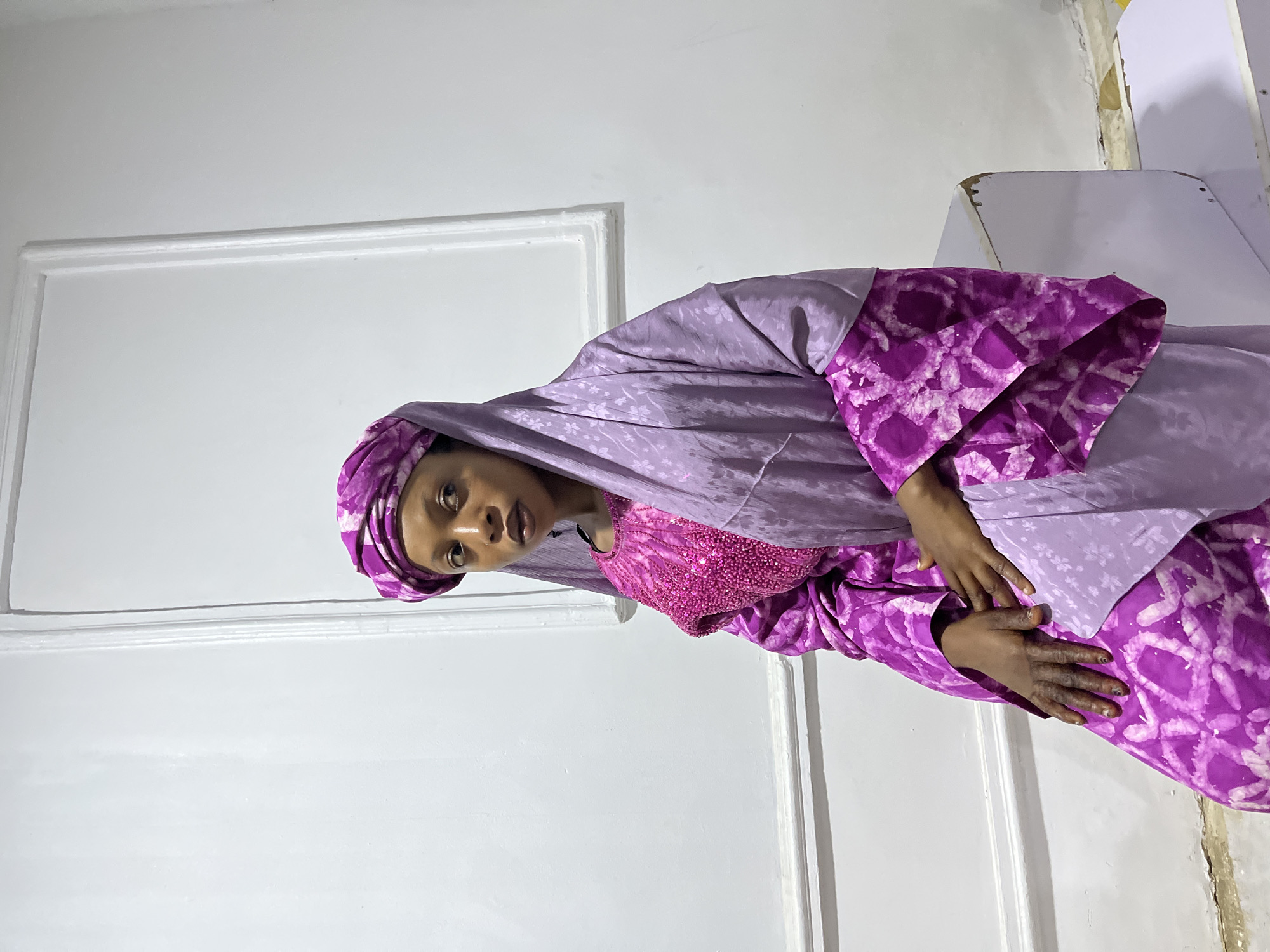 A woman wearing a purple traditional Nigerian outfit, including a head covering, stands indoors against a white wall with a decorative border. She is turned slightly to the right and looking at the viewer. Her hands are clasped together. The location is Gura Top, Nigeria.