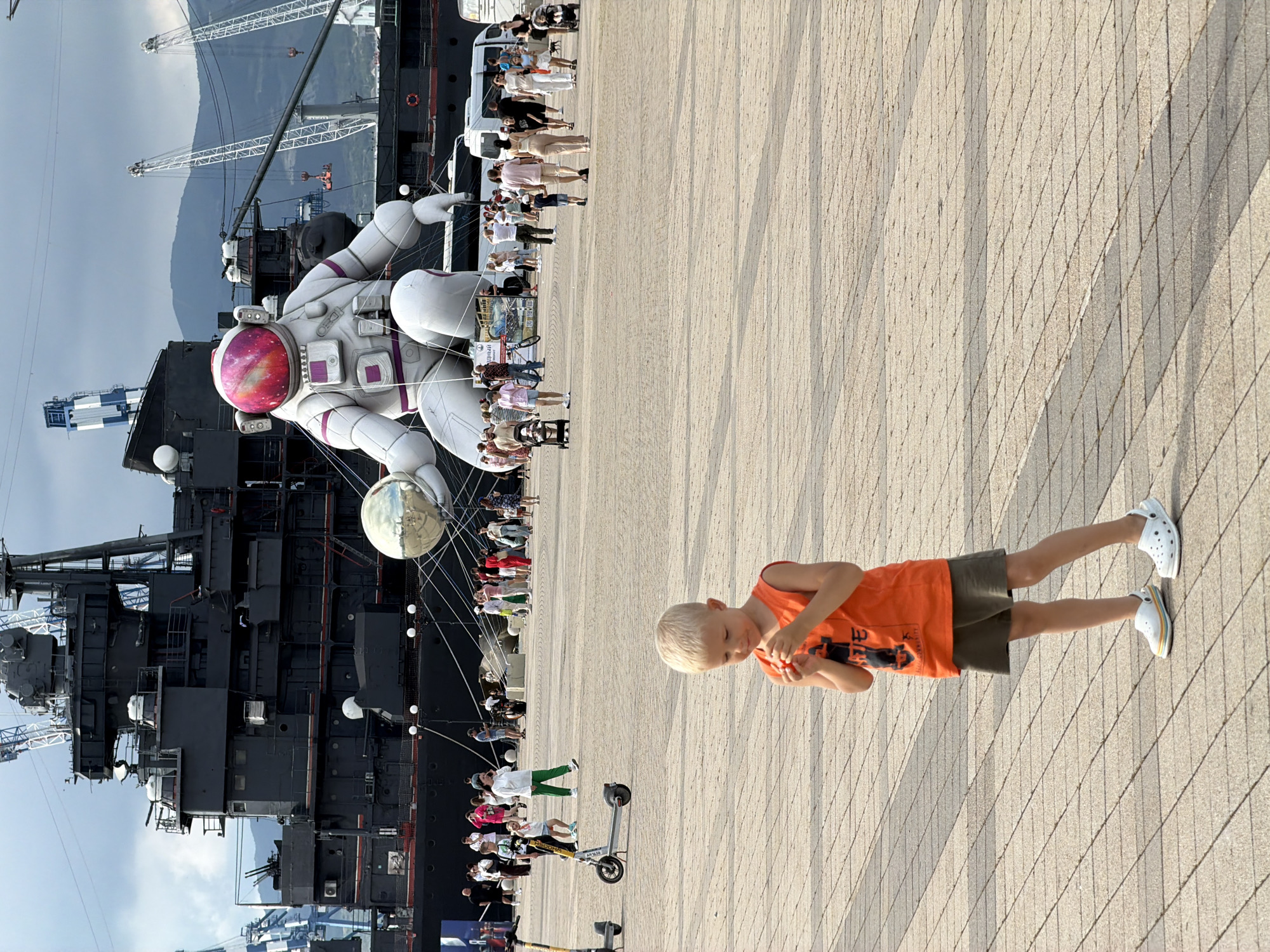A young Caucasian boy with blond hair, wearing an orange sleeveless shirt and olive-green shorts, stands in the foreground on a paved plaza. He is looking down and holding a small red object in his hands. Behind him, a large crowd of people are walking and gathering. In the background, a massive black warship is docked. To the right of the ship, a giant inflatable astronaut figure is prominently displayed, with a reflective sphere in its hands. Several cranes are visible around the ship and the astronaut. The scene appears to be taking place in the port area of Novorossiysk, Russia, with mountains in the distance. The sky is overcast.