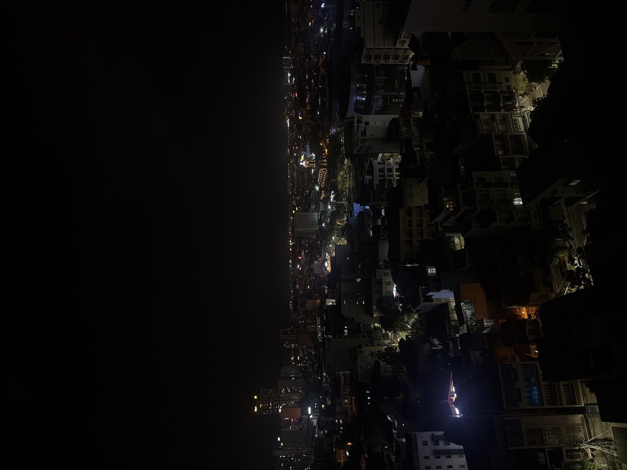 The image shows an aerial view of a city at night, likely Nha Trang Ward, Vietnam, given the context. The right side of the image shows densely packed buildings, with a mix of residential and commercial structures illuminated by streetlights and interior lighting. The structures vary in height, from low-rise buildings to high-rise towers. The left side is predominantly dark, likely representing a body of water or a large, unlit area. The overall light level is low, and the image has a grainy texture, suggesting it was taken in low light conditions. The composition of the image is somewhat tilted, with the horizon appearing to be at an angle.