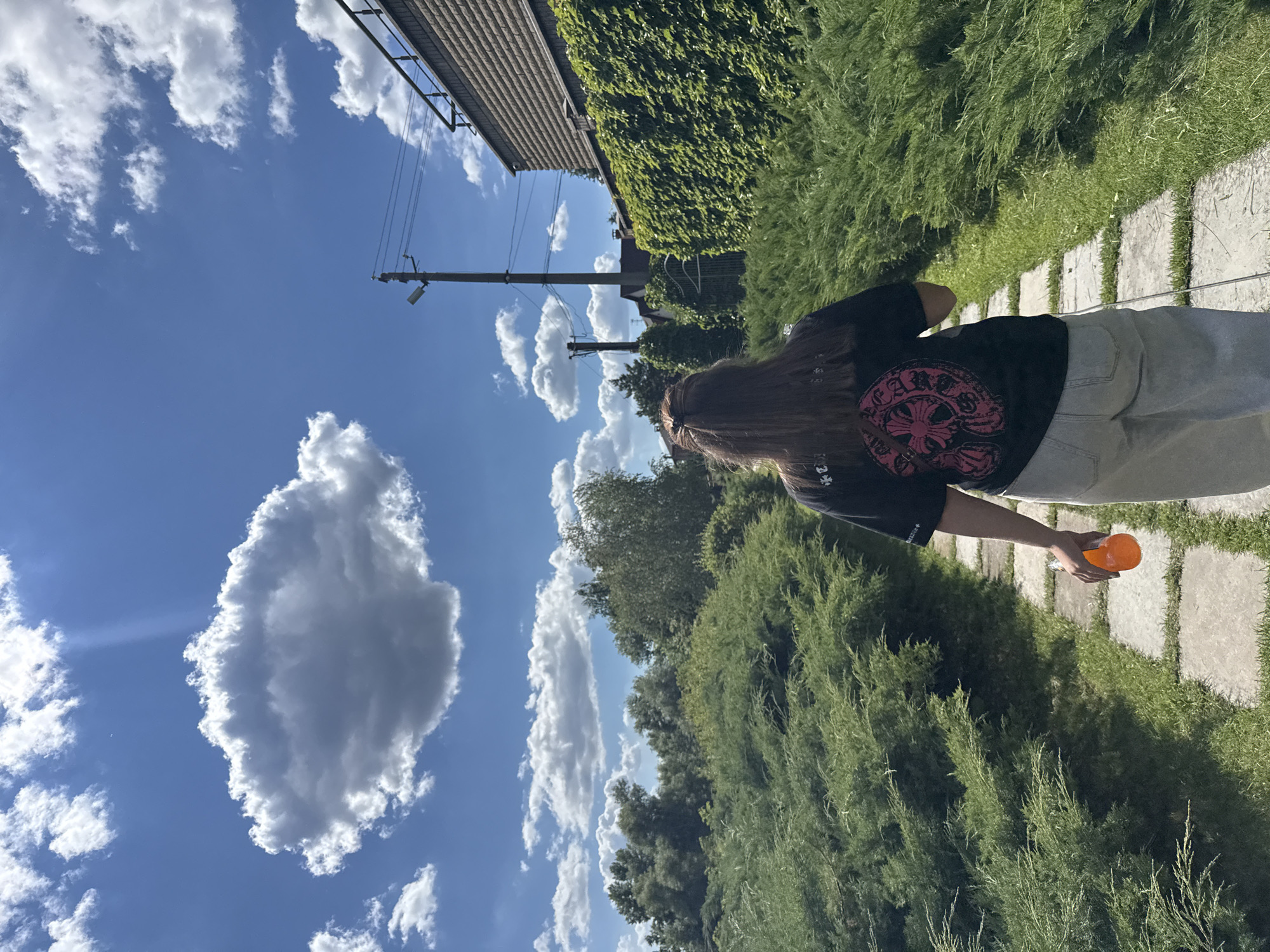 This image shows a person from behind, standing on a paved walkway in what appears to be a garden or park. The person has long, brown hair and is wearing a black t-shirt with a circular pink design on the back and light-colored pants. They are holding an orange object, possibly a bottle or cup, in their right hand.
The setting is lush and green, with dense bushes and trees surrounding the walkway. The sky above is a vibrant blue with several fluffy white clouds. In the background, there is a building with a dark-tiled roof and ivy growing on its side. A utility pole with wires is also visible against the sky.
The lighting suggests it is daytime, likely sunny with some scattered clouds. The overall atmosphere seems relaxed and outdoorsy. The provided location context of Obukhivka, Ukraine, places this scene in a specific geographical area. There is no discernible text in the image.
