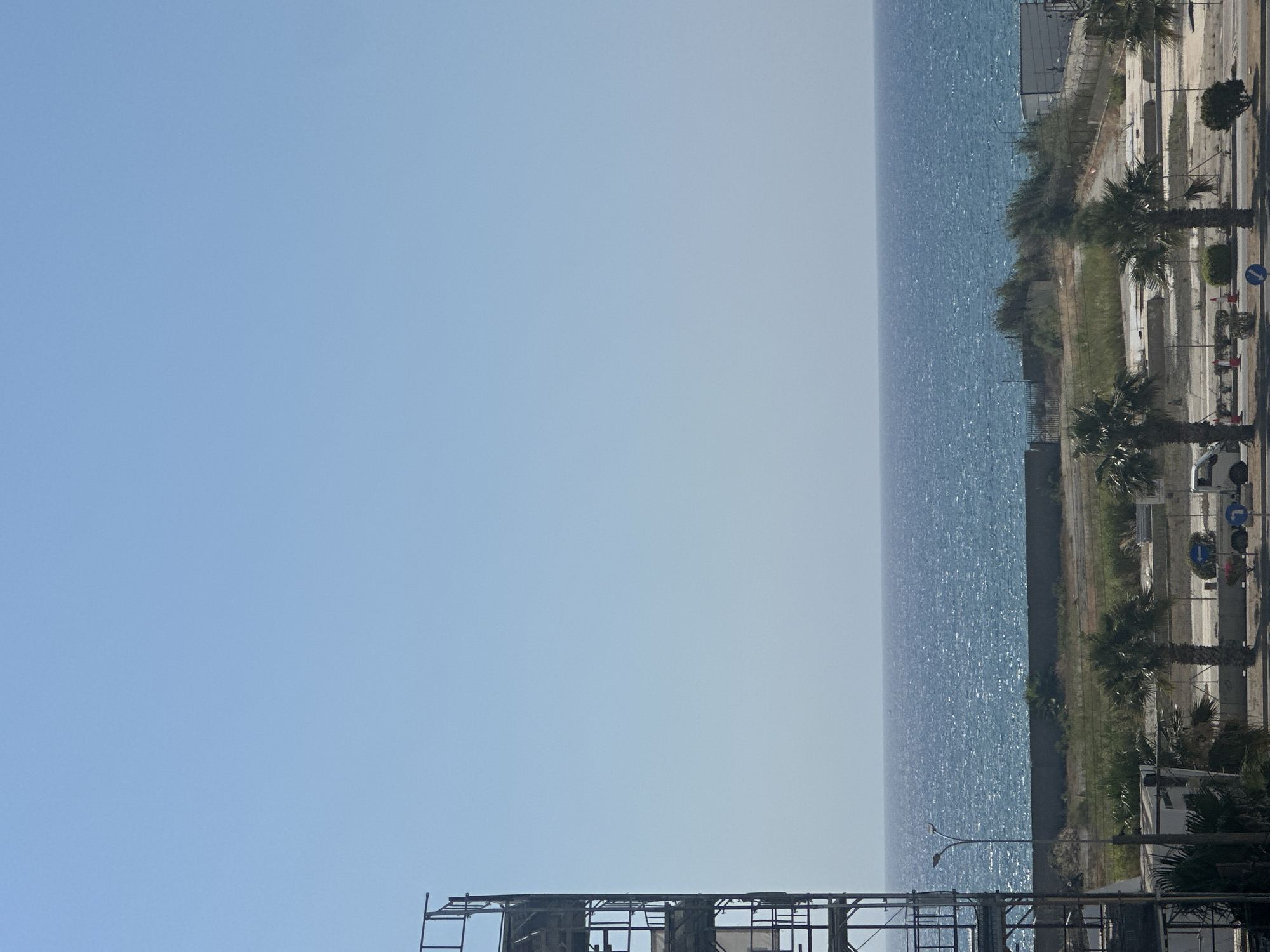 The image depicts a bright, sunny day overlooking the Mediterranean Sea in Larnaca, Cyprus. In the foreground, a partially constructed scaffolding structure stands on the left side of the frame. Below the scaffolding, a white building is visible, partially obscured by palm trees. A street runs horizontally across the lower portion of the image, with traffic cones and road signs indicating construction or road work. A white truck is parked on the street.
Beyond the street and the foreground structures, an open lot with sparse vegetation and some construction debris is present. A low, concrete wall separates this area from the sea. The sea itself stretches across the middle ground, a vast expanse of blue water with a textured surface, indicative of waves or wind. A faint silhouette of a distant boat can be seen on the horizon. The sky above is a clear, pale blue, with no clouds. The overall scene suggests an ongoing construction project near the coastline.