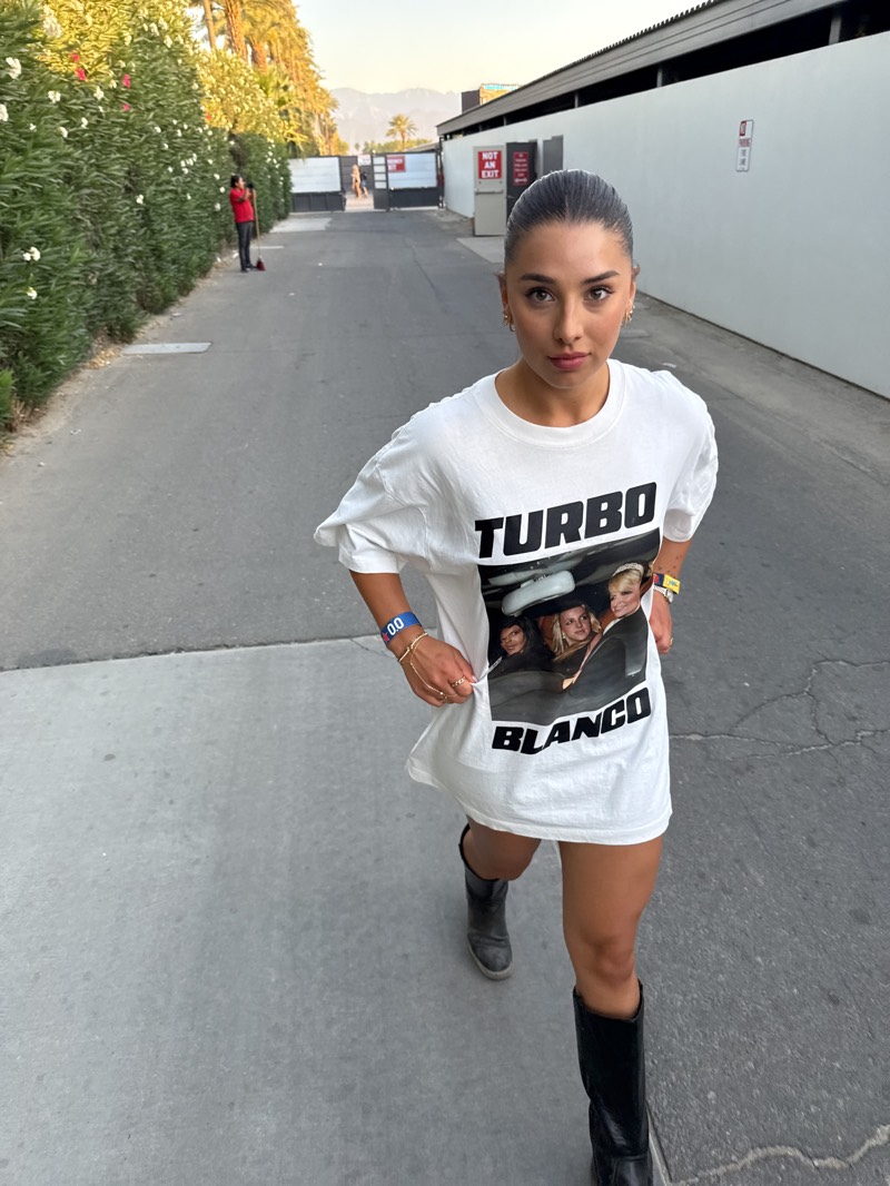 A woman is standing in the city of Indio, United States, wearing a white T-shirt dress with a "Turbo Blanco" graphic, black knee-high boots, and a blue wristband. She is positioned in the middle of a gray paved pathway, looking towards the viewer. The pathway is bordered by tall, dense greenery on the left and a long, white wall on the right with "NOT AN EXIT" sign on it. In the background, a distant gate and mountainous landscape are visible. A person stands on the left behind the greenery, appearing to sweep with a broom.