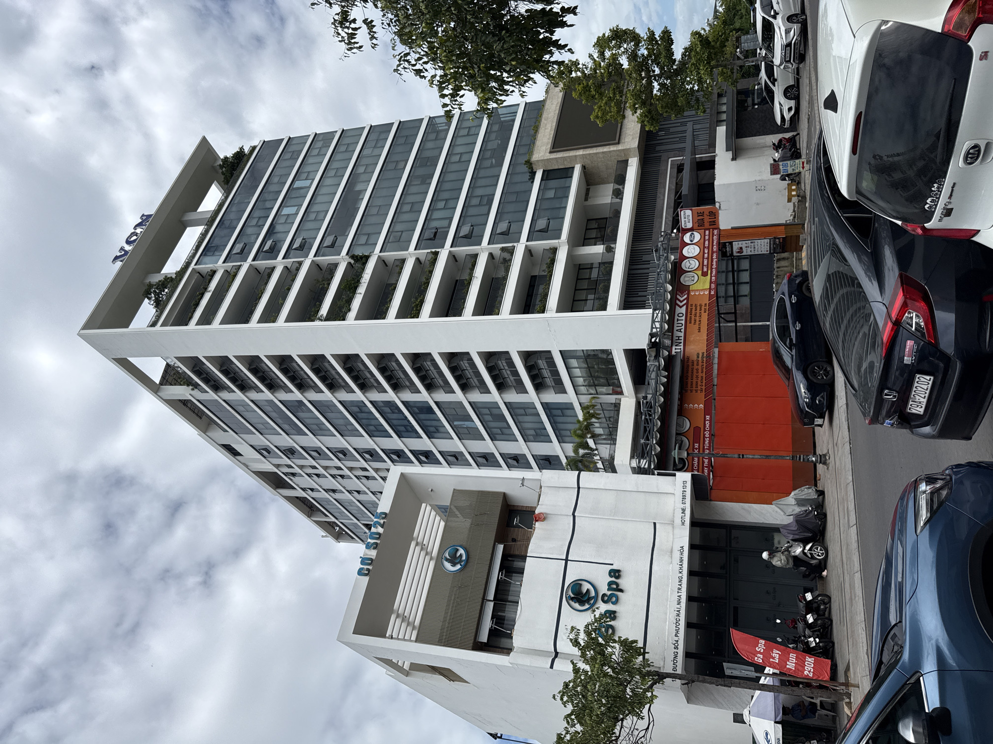The image shows a street view in Nha Trang, Vietnam, dominated by two buildings. The taller building on the right is a modern, multi-story structure with numerous windows and balconies, bearing the "VCN" logo at its apex. To the left, a lower, white building has the signage "Gà Spa" and "CƠ SỞ 25" visible. The street level of the lower building features entrances and windows, with some signs indicating services like "MINH AUTO" and car washing. A red banner with Vietnamese text and "290K" is displayed near a parked scooter. Several cars are parked or driving on the street, including a blue SUV in the foreground, a black sedan, and white sedans further down the road. A person in a light-colored jacket is seen near a parked scooter on the sidewalk. The sky is cloudy, and trees are visible on the right side of the frame. The area appears to be a commercial or mixed-use district.