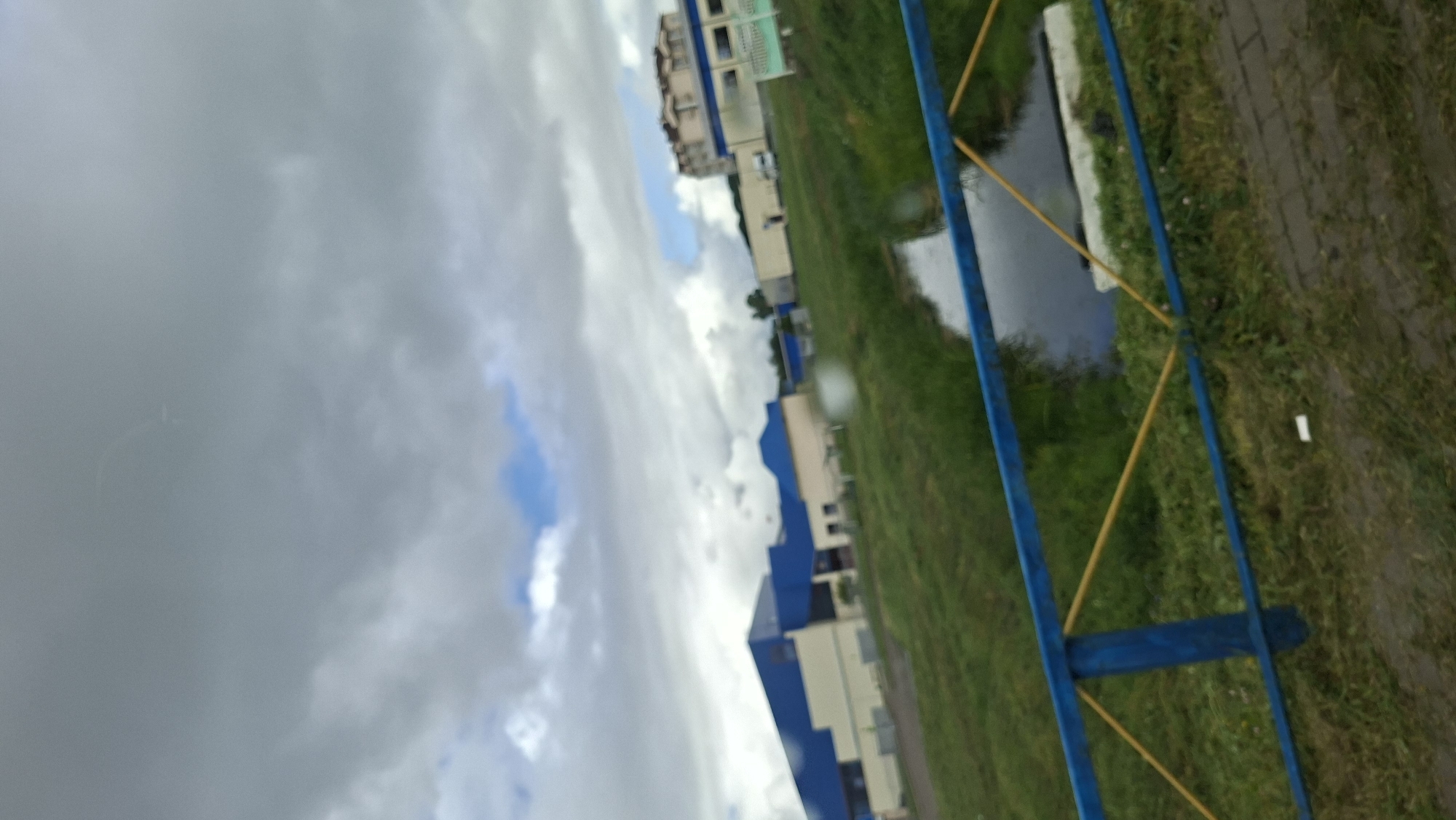 The image shows an outdoor scene in Myadzyel, Belarus, featuring a large building with a blue roof and beige walls, possibly a sports complex or industrial facility. A blue and yellow fence runs across the foreground, partially obscuring a narrow waterway with dark water. The sky is overcast with gray clouds, though a patch of blue sky is visible. The ground is covered in green grass, and there is a paved pathway to the right of the fence. In the background, other buildings and trees are visible. The scene appears to be a public space, likely during daytime.
