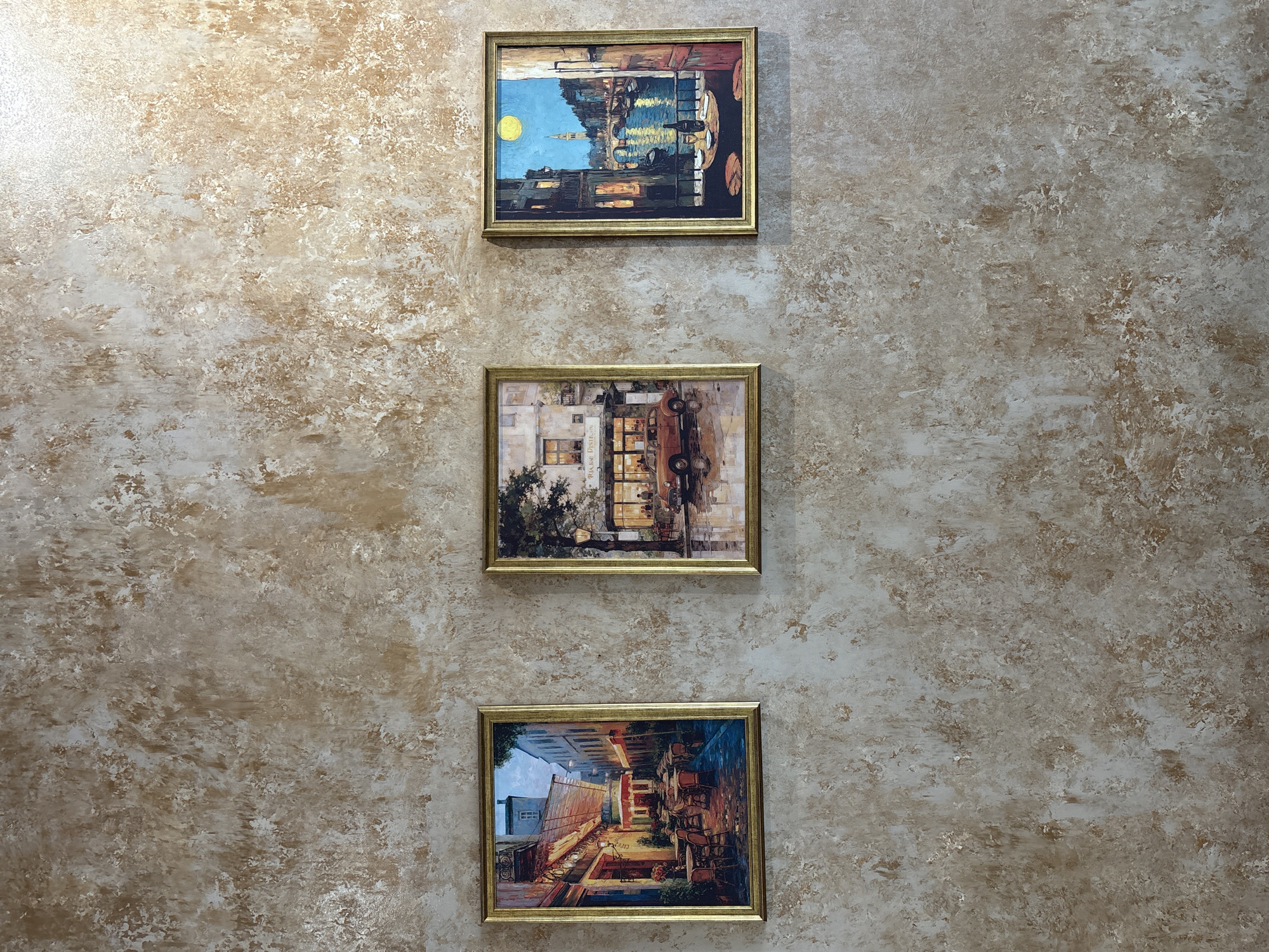 Three framed paintings hang vertically on a textured wall. The top painting depicts a cityscape reflected in water under a bright yellow moon. The middle painting shows a cafe scene with a vintage car. The bottom painting displays a street scene with tables and chairs outside a building. The wall has a mottled beige and white pattern. The precise location is unavailable without further information.