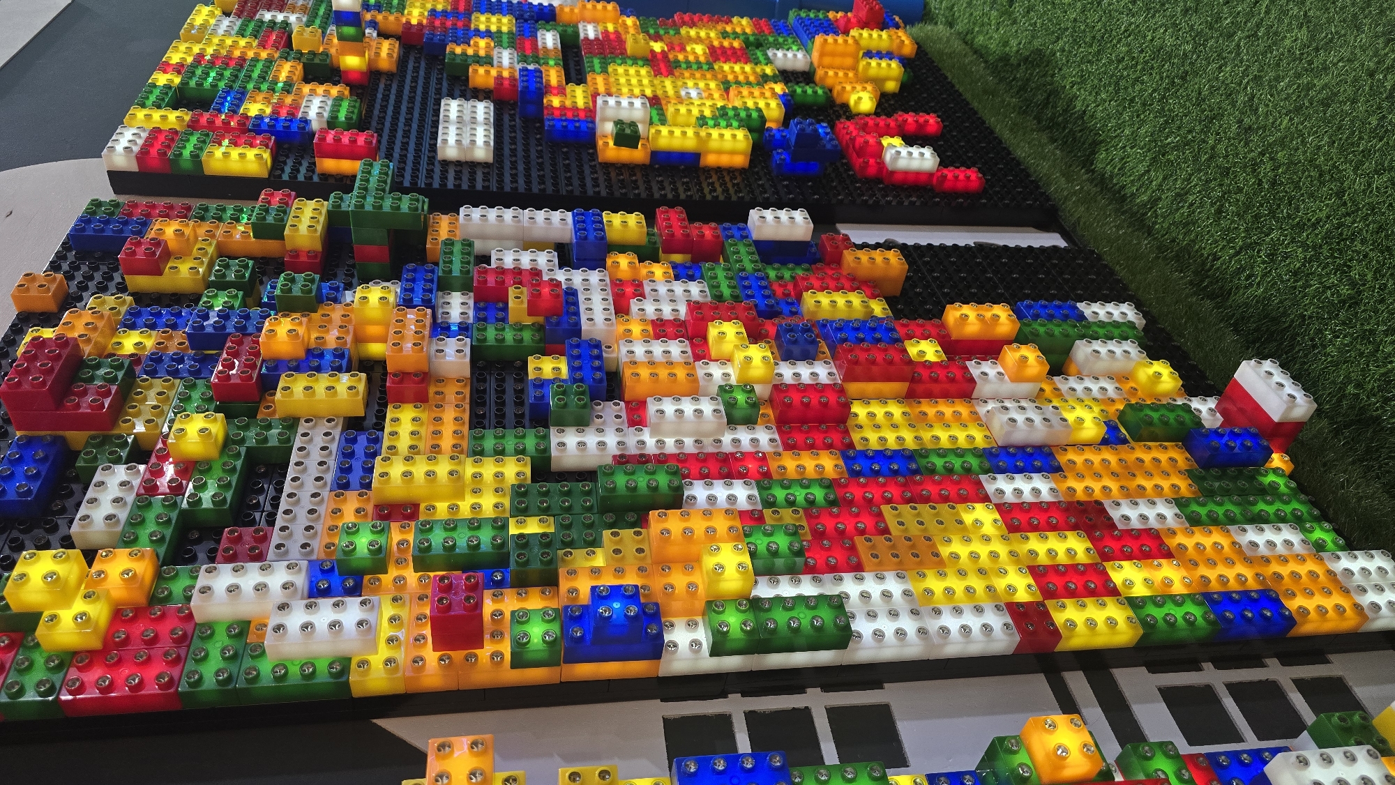 The image displays a vibrant, large-scale wall installation crafted from numerous translucent, internally illuminated blocks, resembling oversized LEGO bricks. The blocks are predominantly red, green, blue, yellow, orange, and white, featuring visible circular studs on their surfaces, which also appear to secure them to a dark, likely black, backing panel. These blocks are arranged in an irregular, three-dimensional relief pattern, with some protruding further than others, creating a textured and uneven surface. Each block emits light from within, contributing to the bright, colorful ambiance of the display. The installation spans across multiple vertical panels. In the foreground and extending towards the right, a bright green artificial turf covers the floor. To the far left, a hint of a light-colored, possibly white, architectural structure with vertical divisions is partially visible, largely obscured by the block display. No individuals are present in the scene. This installation is located in Izhevsk, Russia.