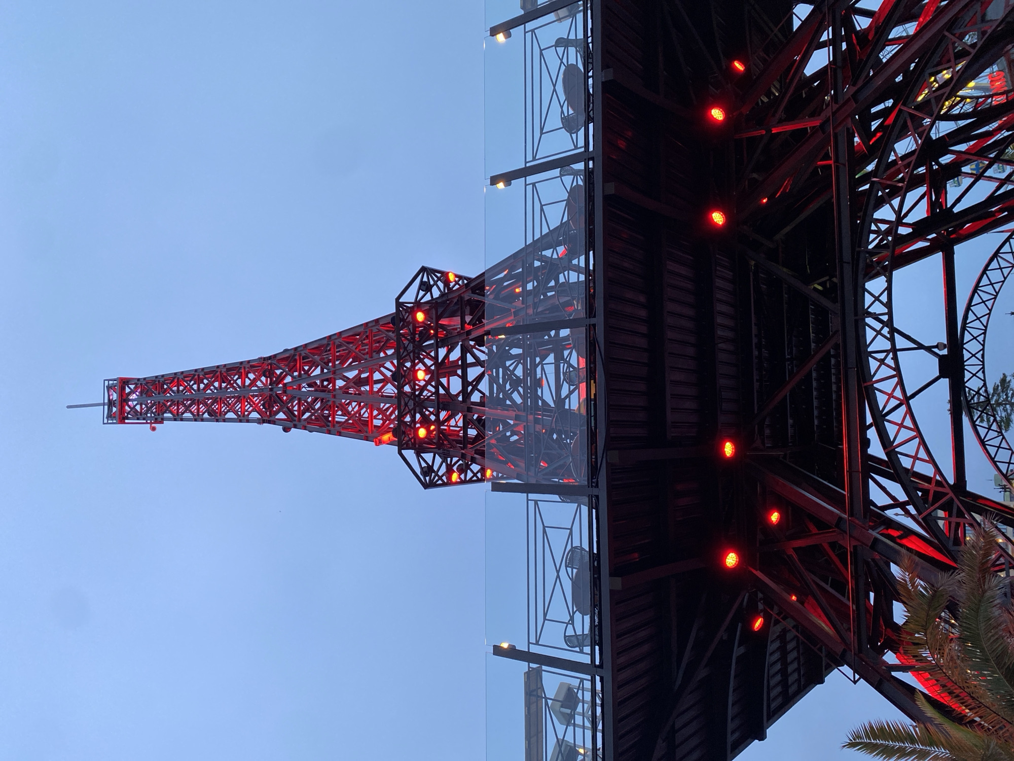 A low-angle, upward-looking shot captures a structure resembling the Eiffel Tower, illuminated with red lights against a clear, pale blue twilight sky in Varna, Bulgaria. The structure is composed of dark metal latticework and beams. Red spotlights cast a glow from within its framework and from beneath an elevated platform.
Approximately one-third up the structure, a viewing or dining platform is visible. This platform features a glass railing with metal posts and horizontal bars. Several dark, woven outdoor chairs are arranged around tables on this platform, suggesting a seating area. Additional, smaller light fixtures are mounted on the railing posts.
The base of the main structure is wide, with multiple diagonal and horizontal beams forming its support. In the bottom left, the fronds of a palm tree are visible, partially lit by the red glow from the structure's underside. Towards the bottom right, through the open framework of the structure, faint lights and parts of what appear to be amusement park rides (possibly a Ferris wheel or roller coaster) are discernible in the distance. The overall scene suggests an evening setting at a recreational or tourist attraction.
