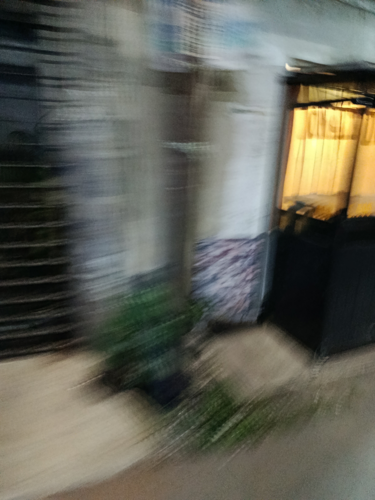 The image shows a blurred scene in Depok, Indonesia, likely at night or in low light conditions, with motion blur. On the left, a dark, striped structure, possibly a fence or shutter, is visible. To the right of this, a lighter, textured wall or surface is present, with a patterned object leaning against it. Further to the right, a window is lit from within, covered by yellow curtains. In front of this window, dark structures, likely furniture or architectural elements, are visible. A green, leafy plant is present in the foreground, also affected by motion blur, suggesting movement. The ground in the foreground appears to be a light-colored surface, possibly a sidewalk or pavement. The overall impression is of a street scene or storefront, with a sense of movement or speed contributing to the blur.