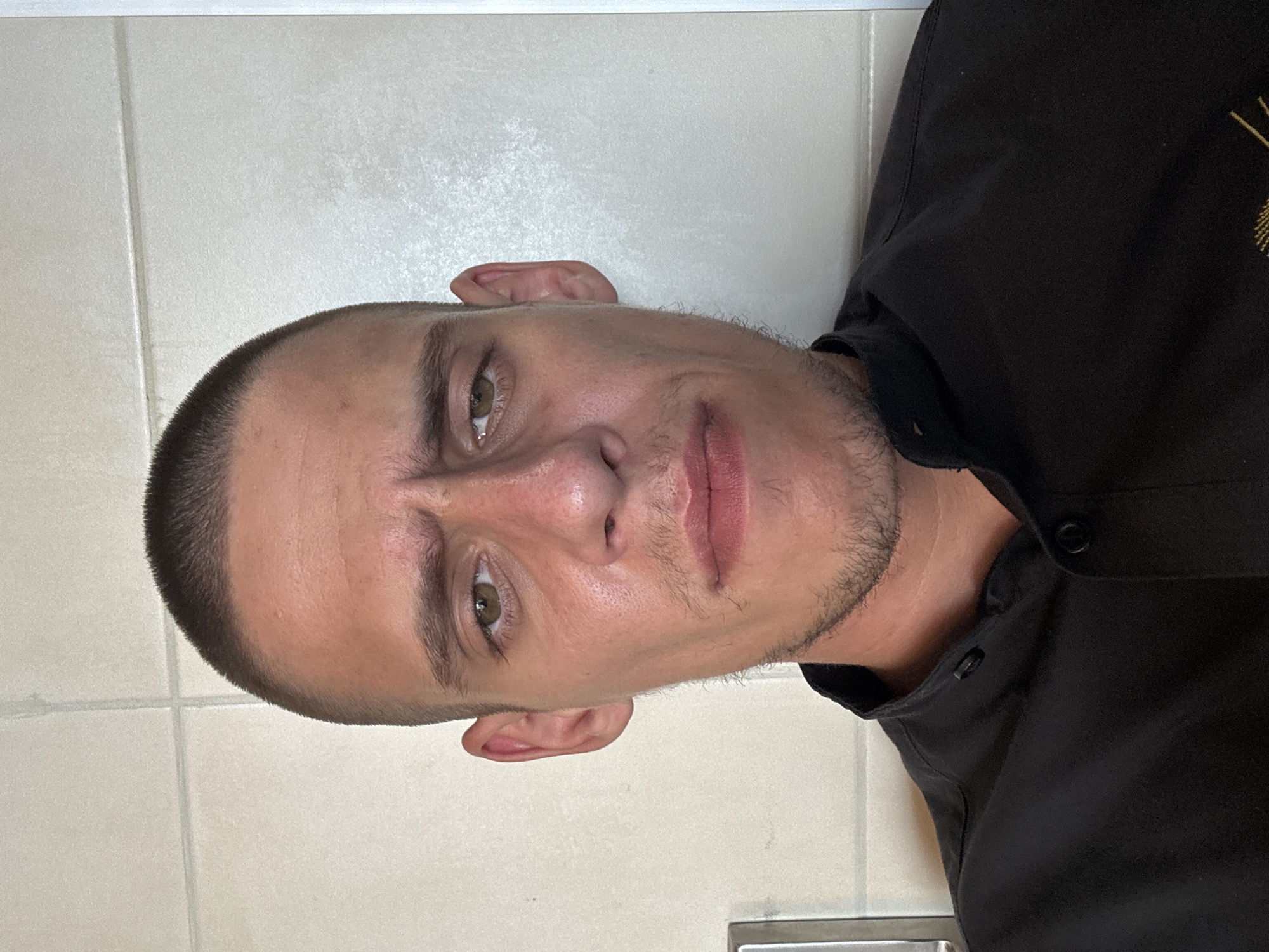 This is a close-up, front-facing selfie of a young adult male with a shaved head and light stubble. He is wearing a black collared shirt. The background consists of tiled walls, suggesting an indoor environment, potentially a bathroom or kitchen. The lighting appears to be from above and slightly to the front, casting subtle shadows. The overall impression is that of a casual, unposed photograph taken at home. There are no identifiable landmarks or indications of the city of Sofia, Bulgaria, in the image.