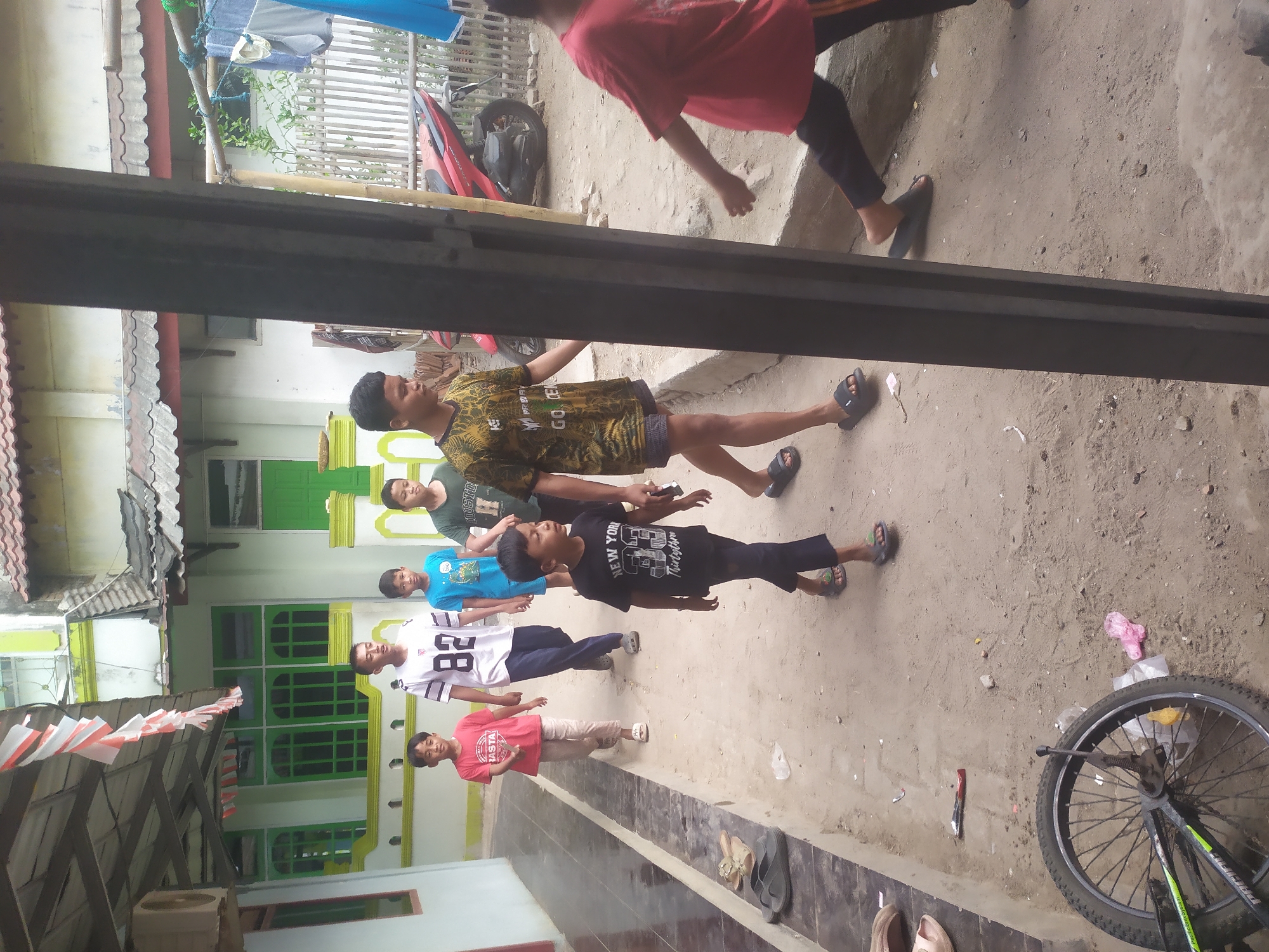 In Kragilan, Indonesia, a group of six young boys walk along a sandy path in front of a house. Two boys in the foreground are walking towards the camera, one wearing a black t-shirt with "New York" printed on it, and another in a yellow and green patterned t-shirt. Behind them, four other boys walk away from the camera. The boy second from the left in this group wears a white t-shirt with the number "82" on it. The building behind the boys has green-framed windows and a red-tiled roof. A red motorcycle is parked in the background. The scene appears to be a residential street.