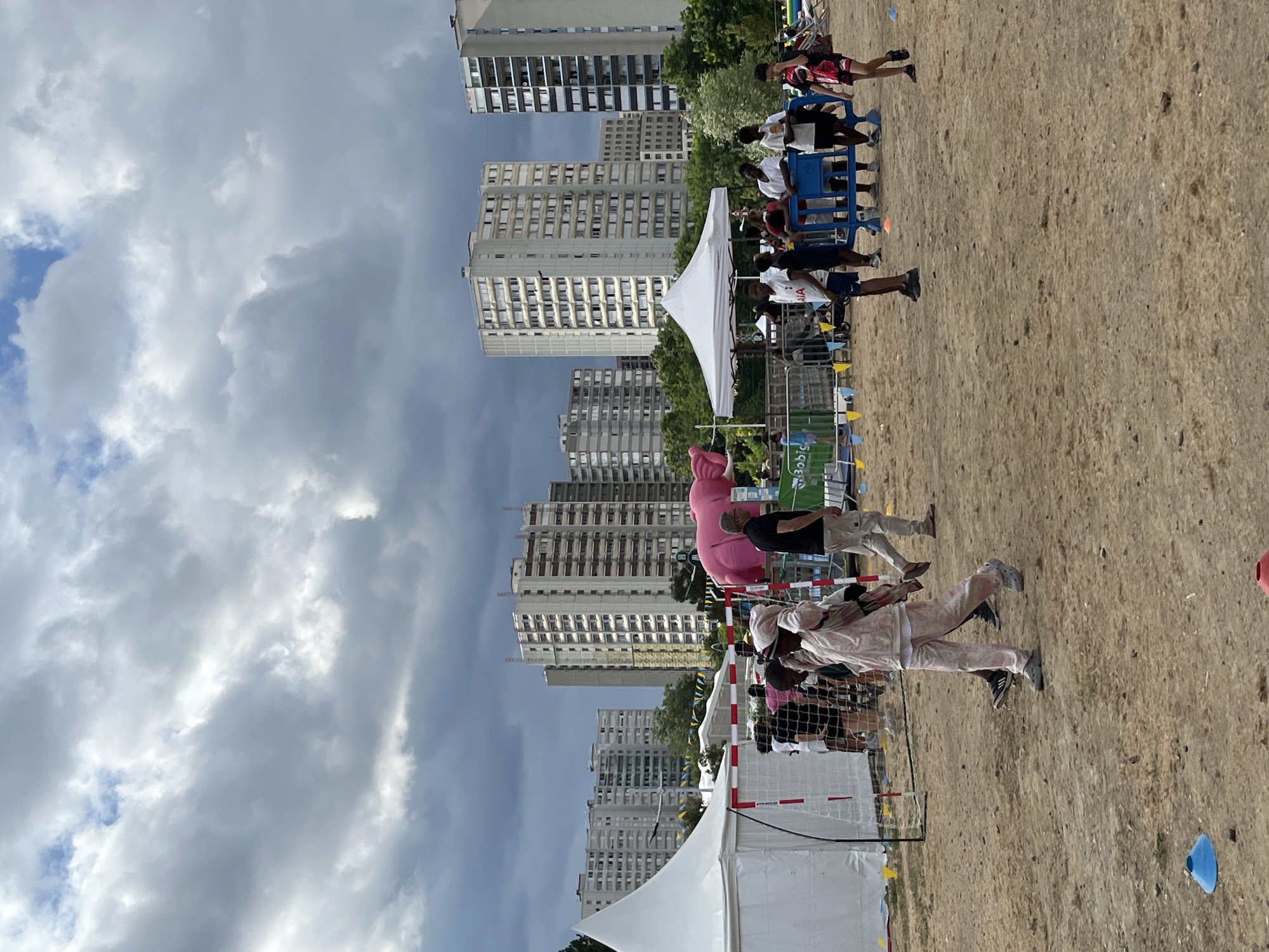 The image depicts an outdoor event on a sandy area in Bobigny, France, under a cloudy sky. Several tall, modern apartment buildings dominate the background. In the foreground, people are gathered around various structures, including white tents and a handball goal. A large, inflatable pink elephant is a prominent, whimsical element. Some individuals are walking, while others appear to be spectating or participating in activities. Children and adults are present, some wearing casual clothing. Cones are scattered on the sand, suggesting a potential sports or game setup. The overall atmosphere suggests a community gathering or festival.