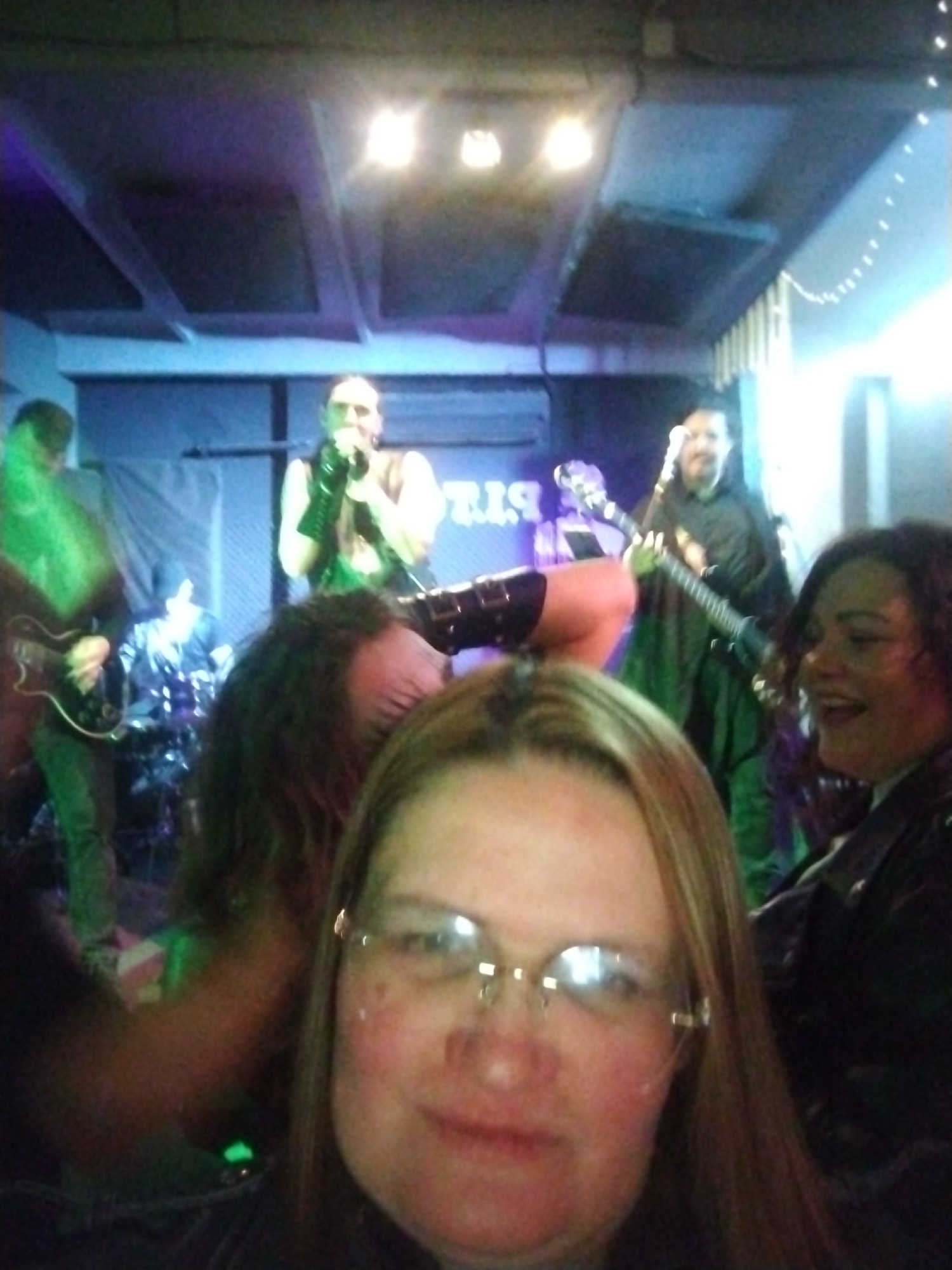 The image appears to be taken at a live music venue, potentially in Chaikovsky, Russia, although this is unconfirmed. Several people are present, with the foreground dominated by a woman with blonde hair and glasses who appears to be taking a selfie. Behind her, a band is performing on a stage. One member of the band is singing, wearing black gloves, while another is playing a guitar. There is at least one other band member visible. The lighting is dim and colored, typical of a concert setting. String lights are visible on the right side of the image.