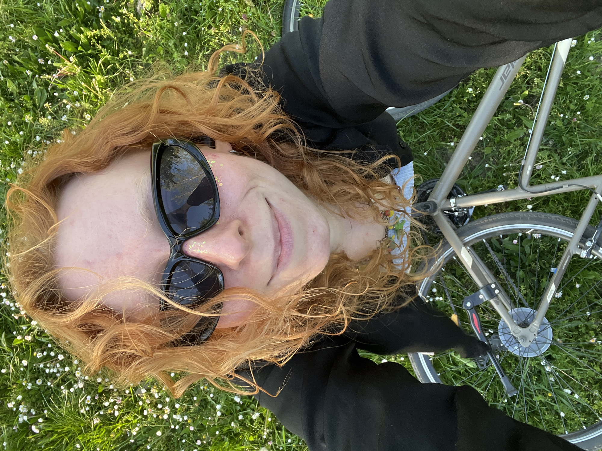 A person is lying on green grass dotted with small white flowers, possibly daisies, in an outdoor location in Cukarica Urban Municipality, Serbia. The person has curly red hair, is wearing black sunglasses with gold glitter on the frames, and is dressed in a black top. A portion of a silver bicycle is visible to the right of the person, including a wheel, part of the frame, and a kickstand. The person is smiling slightly.