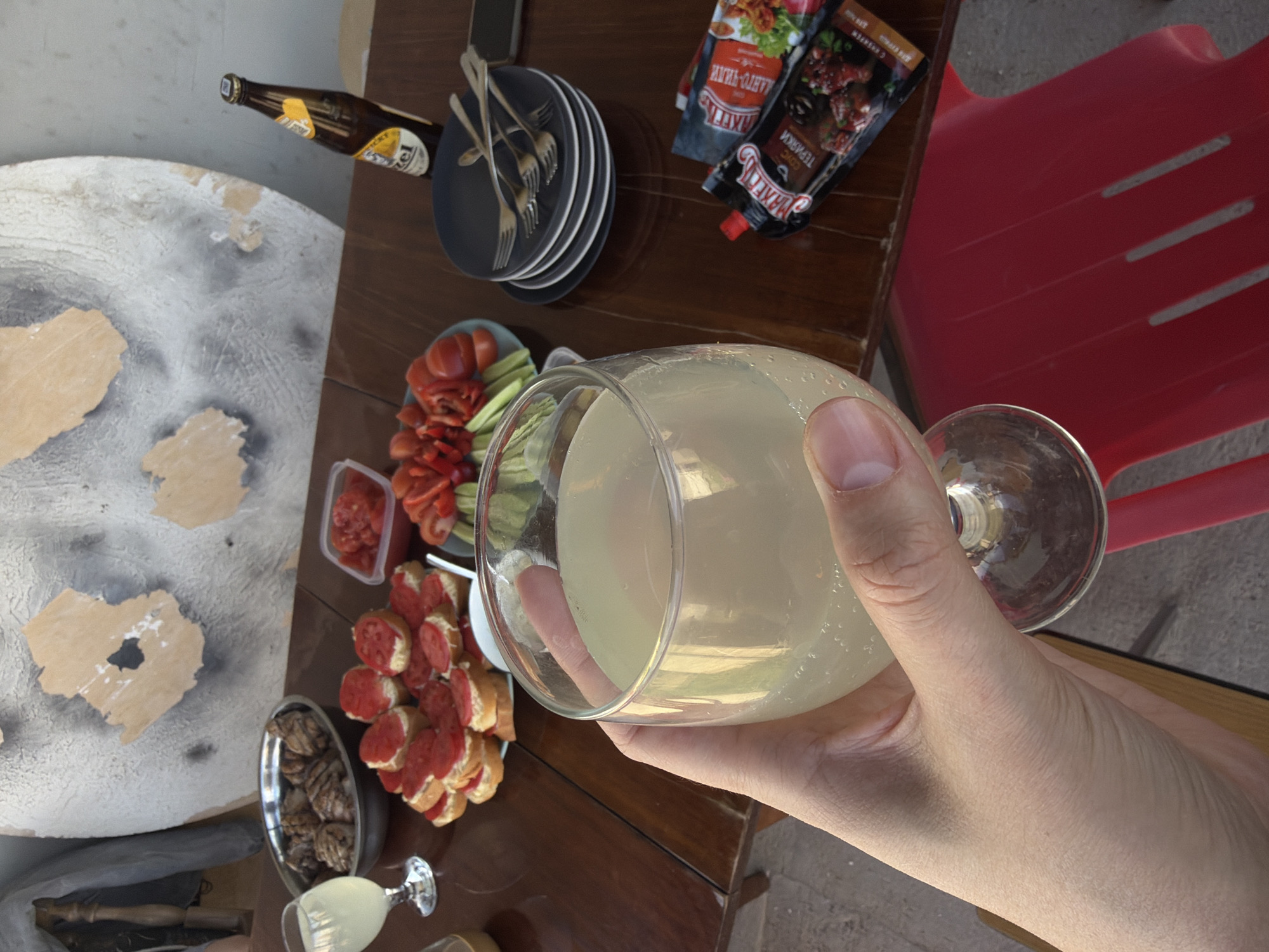 The image depicts a table laden with food and drink, including sliced tomatoes and cucumbers, sandwiches with sliced sausage, nuts in a bowl, a bottle of beer, and condiments in packets. A stack of plates and cutlery are also visible. A hand holds a large glass filled with a light-colored beverage. A red plastic chair is partially visible in the background. A round table with a textured surface mimicking a map or geological feature is situated to the left. The image was likely taken in Chaikovsky, Russia, based on visual clues and the user's prompt.