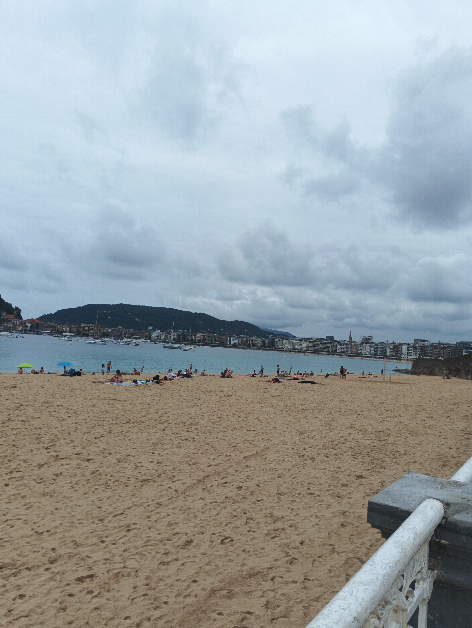 This is a wide shot of a sandy beach on a cloudy day. In the foreground, the sand is marked with footprints. Towards the middle ground, several people are spread out on the sand, some lying down and others sitting. Two beach umbrellas are visible, one green and one blue. In the background, the ocean stretches out, dotted with several sailboats. A city with buildings and a hill covered in greenery rises behind the bay. The sky is filled with dramatic, gray clouds.