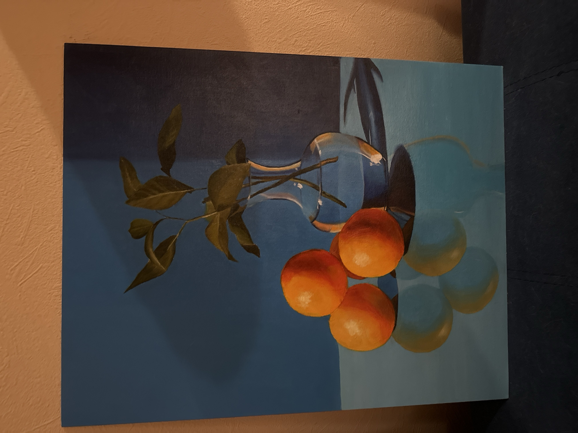 The image is a still life painting. It features a clear glass vase lying on its side with stems and green leaves extending from its opening. Four bright orange fruits are arranged near the vase's opening, with their translucent blue shadows beneath them. The background of the painting is a two-toned blue, with a darker shade on the left and a lighter shade on the right, divided by a vertical line behind the vase. The painting is set on a wall next to another wall with beige finish. Additionally, there is a dark blue material visible on the right edge of the painting. The location is not discernible from the image.