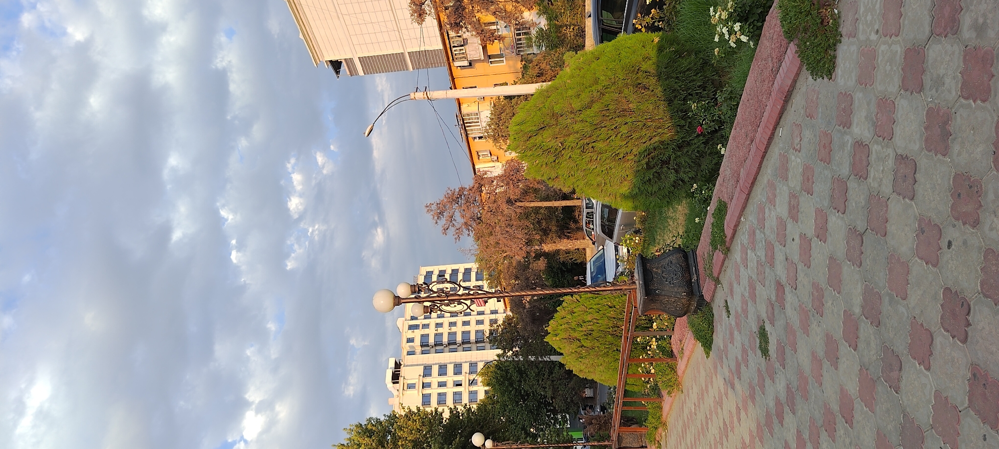 The image captures a view of a street in Khujand, Tajikistan, on a cloudy day. In the foreground, a patterned sidewalk made of interlocking red and gray tiles leads into a park-like area with manicured green bushes. A decorative black planter sits near the bushes, and a metal fence is visible. Streetlights with white spherical lamps are present, one in the center and another further to the left.
In the background, several buildings are visible. A tall, modern white building with many blue-tinted windows stands prominently on the left. To its right, an orange building with balconies and various windows is situated. Further to the right, a larger white building with a darker, striped section on its side is visible. Several cars are parked along the street. The sky is filled with a mix of white and gray clouds, suggesting an overcast or partly cloudy condition. Trees with green and some reddish-brown foliage are scattered throughout the scene. The overall impression is an urban streetscape with elements of landscaping and residential/commercial buildings.