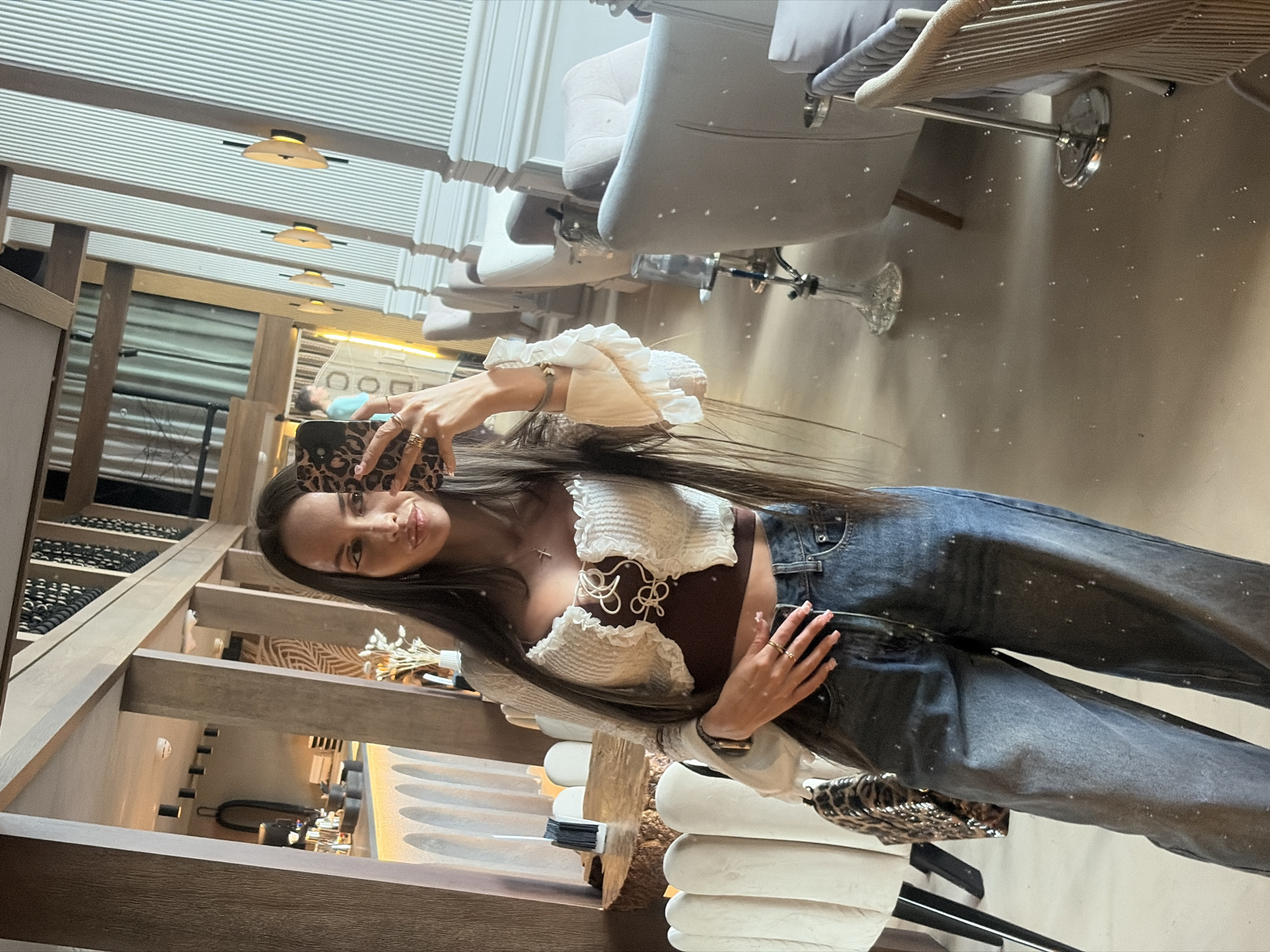 A full-body, slightly angled, mirror selfie shows a young woman with long dark hair and a white crop top with ruffled sleeves and a brown bustier underneath. She is wearing wide-leg, distressed denim jeans. She is holding a leopard-print phone in her right hand, partially obscuring her face. In the background, the interior of a restaurant or lounge is visible, with elegant seating areas, curved architectural elements, and decorative lighting fixtures. The environment appears to be clean and well-lit, with a neutral color palette. The scene suggests a casual outing in a modern urban setting. The presence of hookah pipes and plush seating indicates a lounge atmosphere, potentially in a high-end establishment. The decor suggests a contemporary European aesthetic, possibly aligning with the trendy dining and nightlife scene found in cities like Saint Petersburg, Russia.