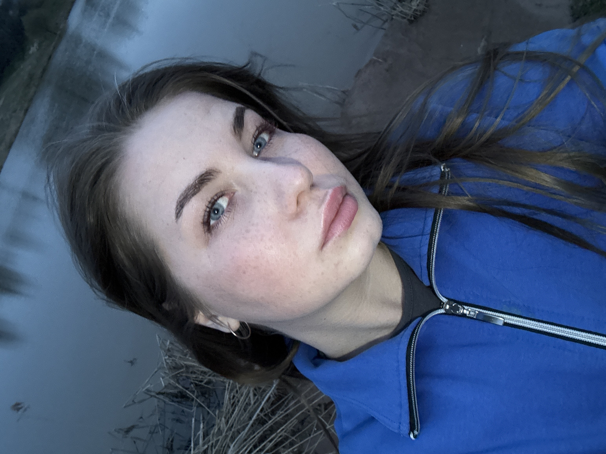 This close-up portrait, taken in Ol'khovochka, Russia, features a young woman with fair skin, prominent freckles across her nose and cheeks, and vivid blue eyes. Her dark brown hair is styled casually, and she wears a small hoop earring in her visible left ear. Her lips are full and appear to have a light gloss. She is dressed in a bright blue zip-up jacket with a distinctive black and white zipper, worn over a dark grey or black inner layer.
The setting is outdoors, with a tranquil body of water, likely a lake or river, forming the background. Dry, light-colored reeds or tall grasses are visible along the water's edge and in the foreground, suggesting a natural, possibly rural environment. The lighting is soft and diffused, creating a cool, subdued atmosphere, which could indicate either an overcast day, early morning, or dusk. The woman maintains a direct, neutral gaze towards the camera. There is no visible text in the media file.