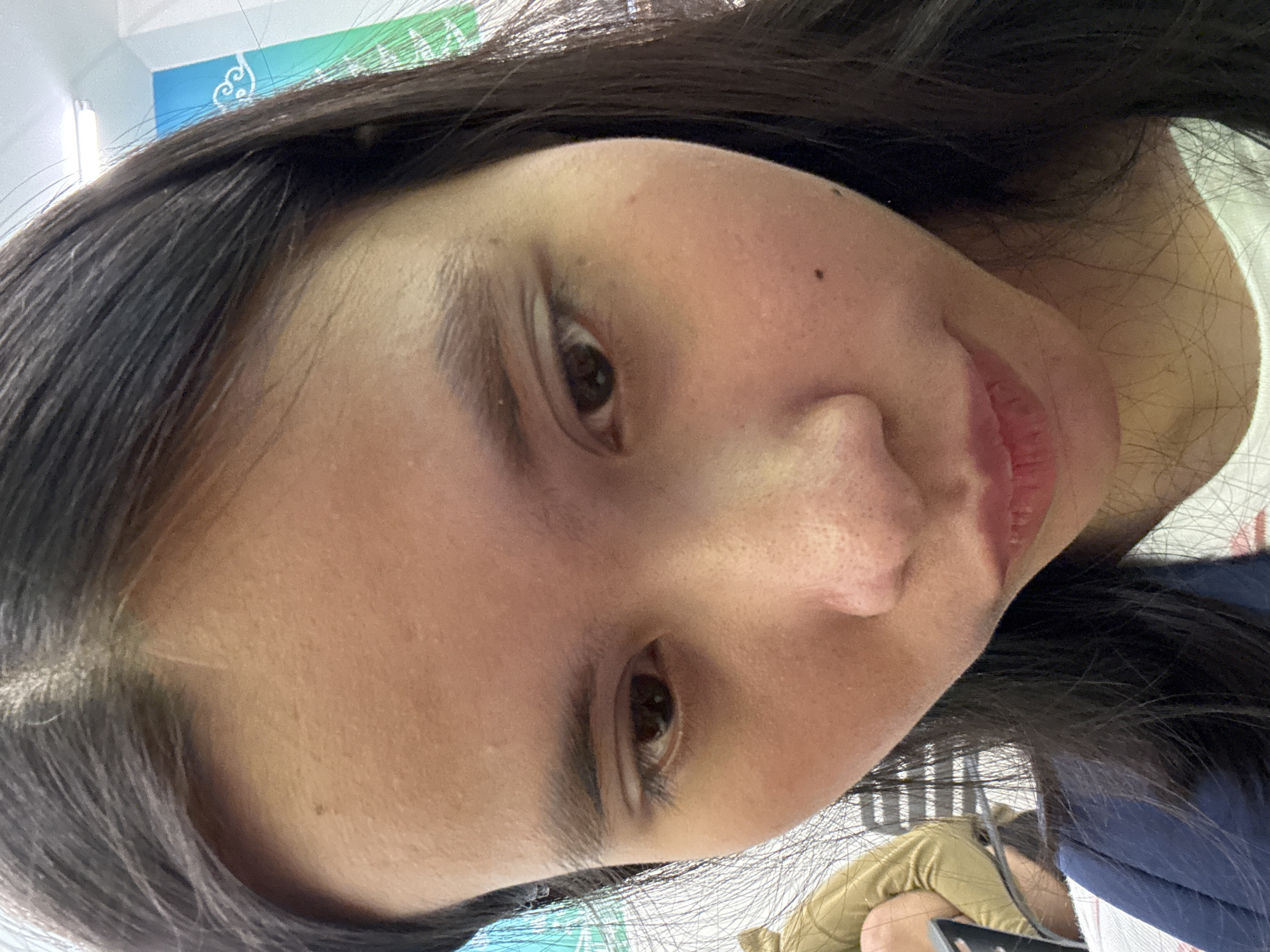 The image is a close-up, low-angle selfie of a young East Asian woman. She has dark hair and is looking down. Her face is the primary focus, with visible pores on her nose and cheeks, and a small mole on her right cheek. She is wearing a white shirt with a blue collar. In the background, out of focus, is a person wearing a light brown jacket and a wall with a blue and green banner. The scene appears to be indoors, possibly in an office or public space, given the banner and lighting. The environment suggests a setting within East Jakarta, Indonesia.