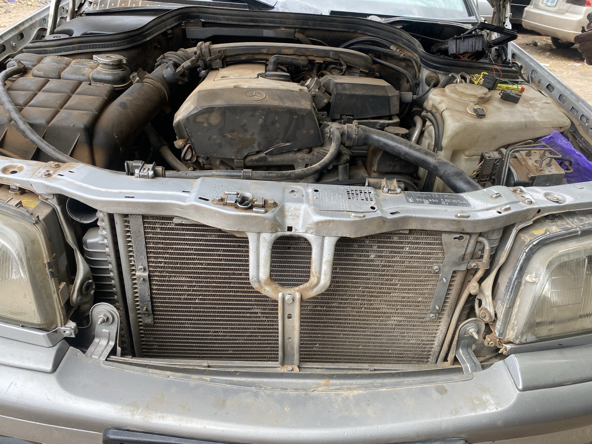 The image shows the engine compartment of a silver Mercedes-Benz car. The engine is covered in dust and grime, indicating it may have been sitting for a while or is used in a dusty environment. Several components are visible, including the engine block, hoses, wires, and a radiator. The car appears to be parked outdoors in what seems to be a workshop or a similar setting. In the background, parts of other vehicles and a person's hand are visible, suggesting some activity is taking place around the car. The overall impression is one of mechanical work or maintenance being performed on the vehicle. The scene is likely located in Jalingo, Nigeria, given the context provided.