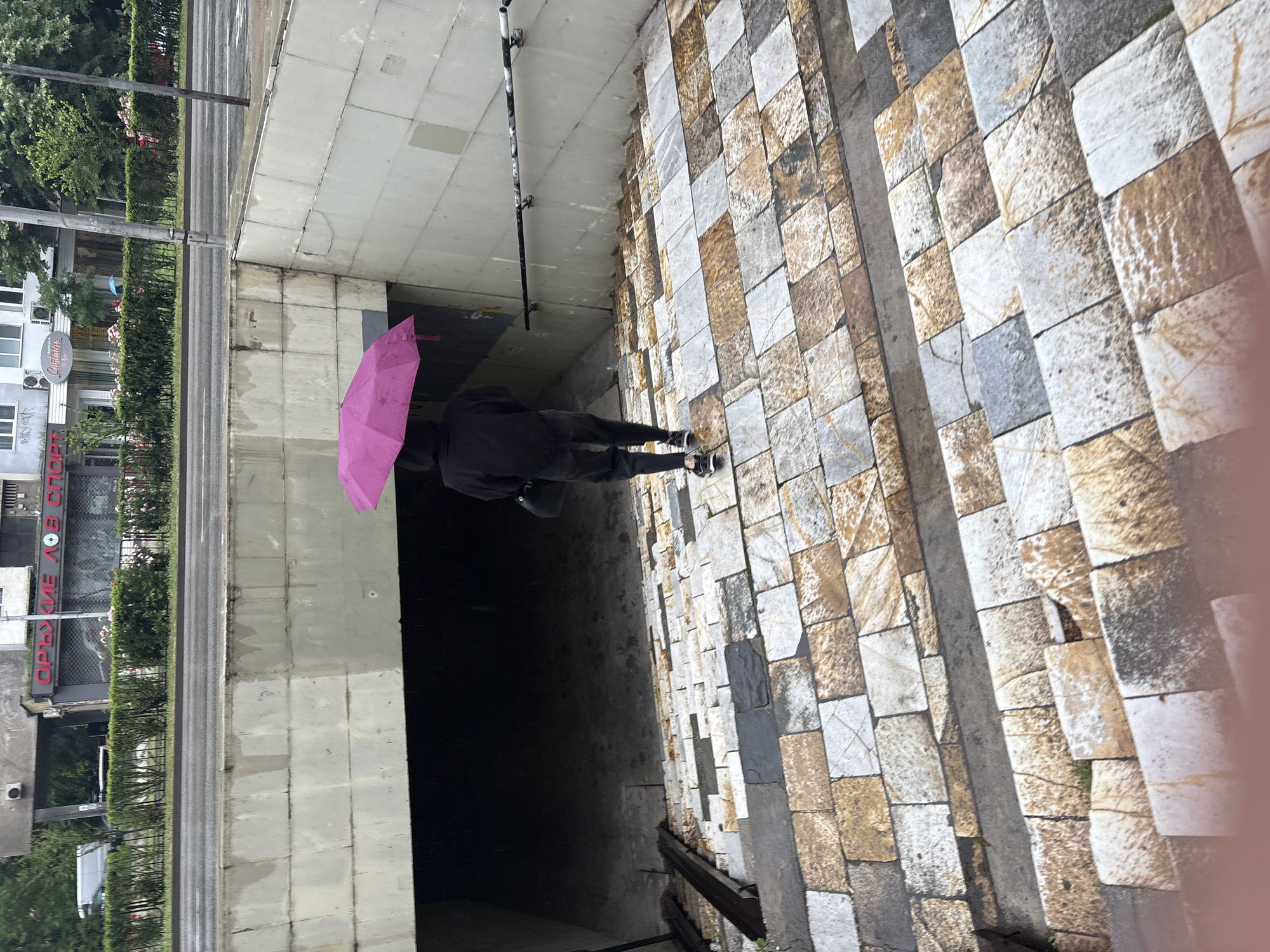 An eye-level photograph depicts a person with a pink umbrella entering a dark tunnel. The individual is dressed in dark clothing. The tunnel walls are constructed of light gray concrete blocks. The pavement is made of cobblestones in mixed brown and gray shades. In the background, buildings and greenery are visible. The photograph was taken in Plovdiv, Bulgaria.