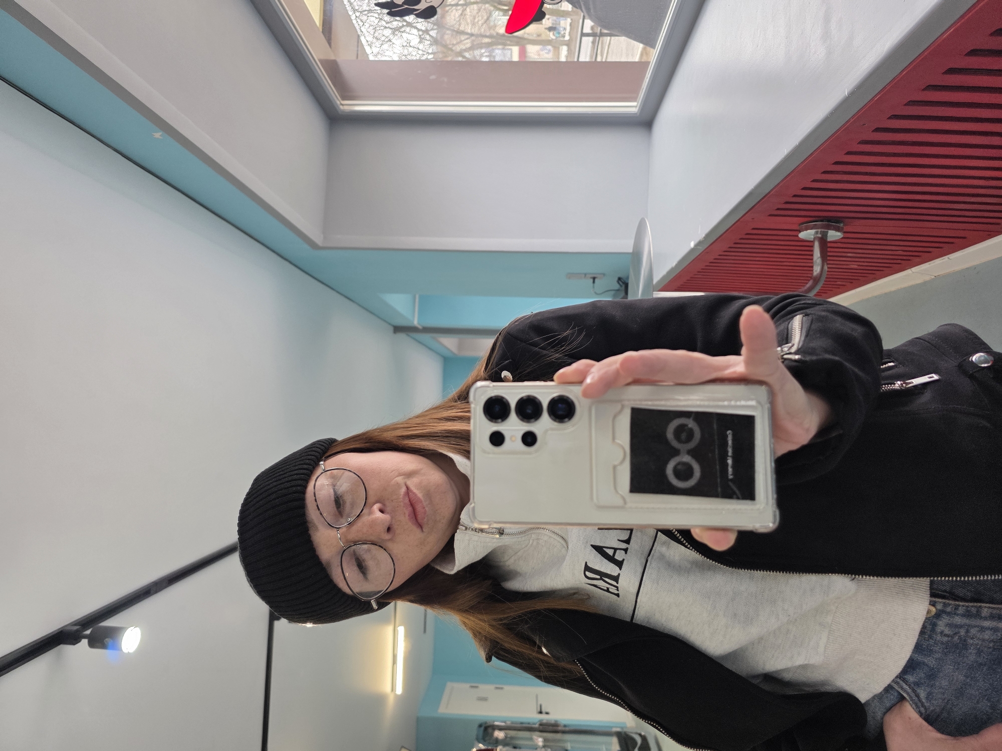 A person is holding a phone, taking a selfie in what appears to be an interior space with a red and grey ceiling and blue walls. The person is wearing a black beanie, glasses, a grey sweater, and a black jacket. The phone is in a transparent case. The image shows a partial view of a window and an overhead lighting fixture. The location, Chaikovsky, Russia, cannot be confirmed based on visual information.