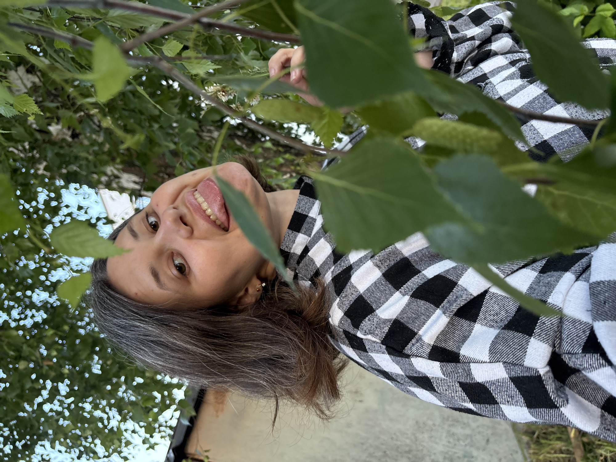 A young woman with shoulder-length dark hair, streaked with grey, is photographed from the chest up, looking towards the camera. She is wearing a black and white checkered shirt. Her head is tilted, and her tongue is sticking out, with her mouth open. Her left ear is visible, adorned with a small stud earring. The background consists of blurred green foliage and the side of a building with a pale, textured wall. The lighting suggests an outdoor setting, possibly in a residential area. The woman appears to be standing outdoors, partially obscured by tree branches and leaves in the foreground.