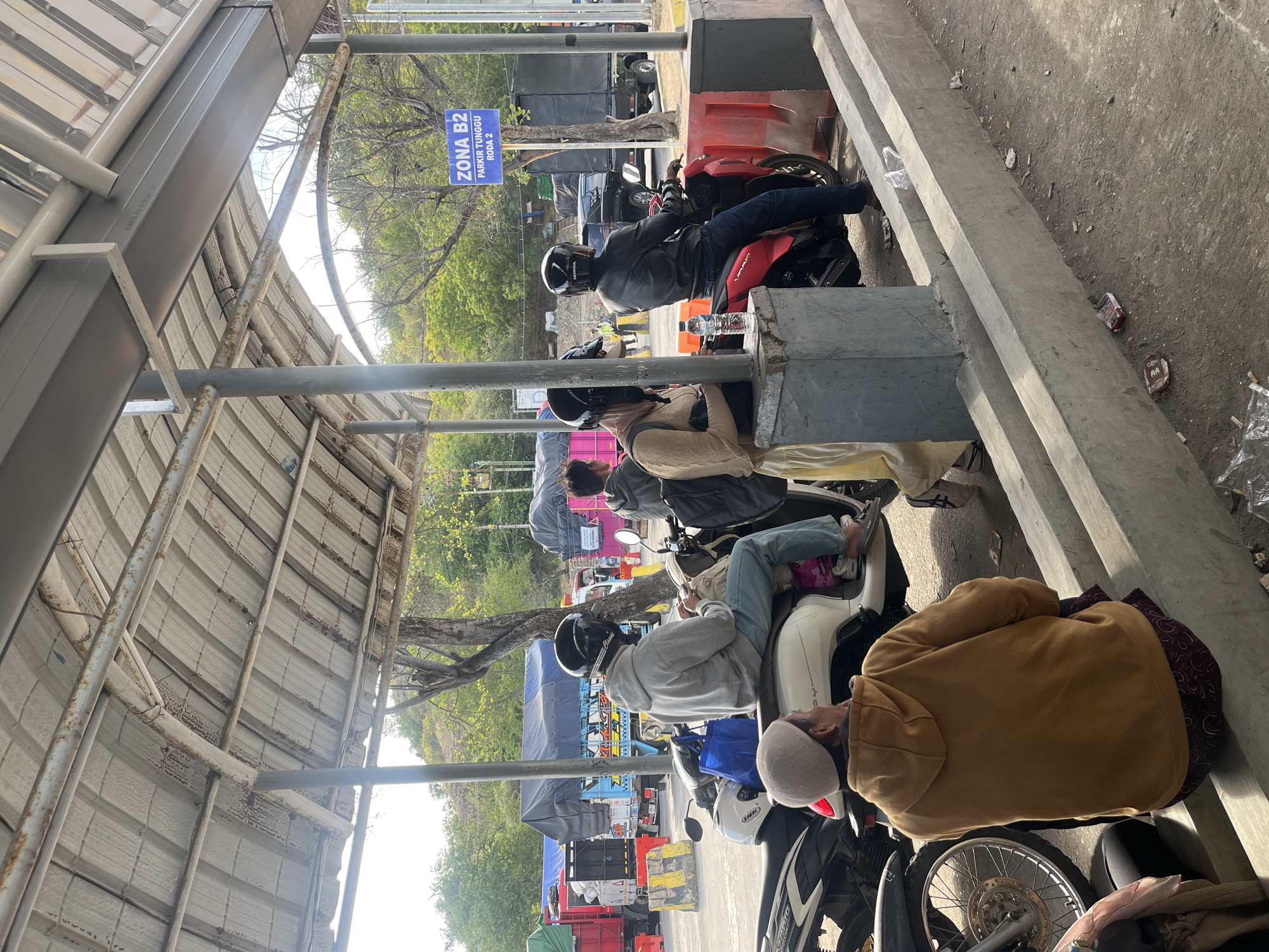 The image depicts an outdoor scene, likely a parking or waiting area, under a covered structure. Several people are present, some seated on motorcycles and others standing. In the foreground, a person wearing a brown hooded sweatshirt sits on a low concrete barrier, facing away from the camera. To their left, the wheel and front of a black motorcycle are visible. To the right, two individuals are seated on white and red motorcycles, respectively. The person on the white motorcycle wears a grey hoodie and blue jeans, while the person on the red motorcycle wears a black leather jacket and blue jeans. Both are wearing helmets. Further back, other individuals and several trucks, some with tarpaulin covers, are visible, suggesting a transportation hub or a point of transit. A blue sign with white text stating "ZONA B2 PARKIR TUNGGU RODA 2" is mounted on a pole to the right. The environment appears somewhat weathered, with exposed metal structures and concrete barriers. The ground is paved and shows signs of debris. The overall impression is one of a temporary waiting area, possibly at a ferry terminal or a border crossing, given the presence of trucks and multiple individuals on motorcycles. The context of Upt Tambak Sari, Indonesia, suggests this is a location within that administrative area, likely a public facility.