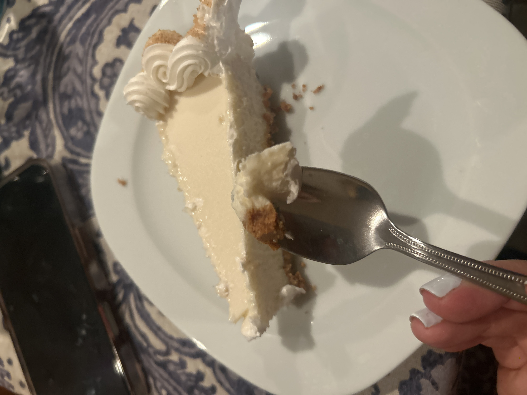 A high-angle shot shows a slice of cheesecake on a white plate, with a portion being lifted by a silver spoon held by a hand with white painted fingernails. The cheesecake has a light-colored filling, a crust, and is topped with swirls of whipped cream. The plate sits on a surface with a blue and white patterned fabric. A black electronic device, possibly a smartphone, is visible on the left side of the frame. The location is unknown.