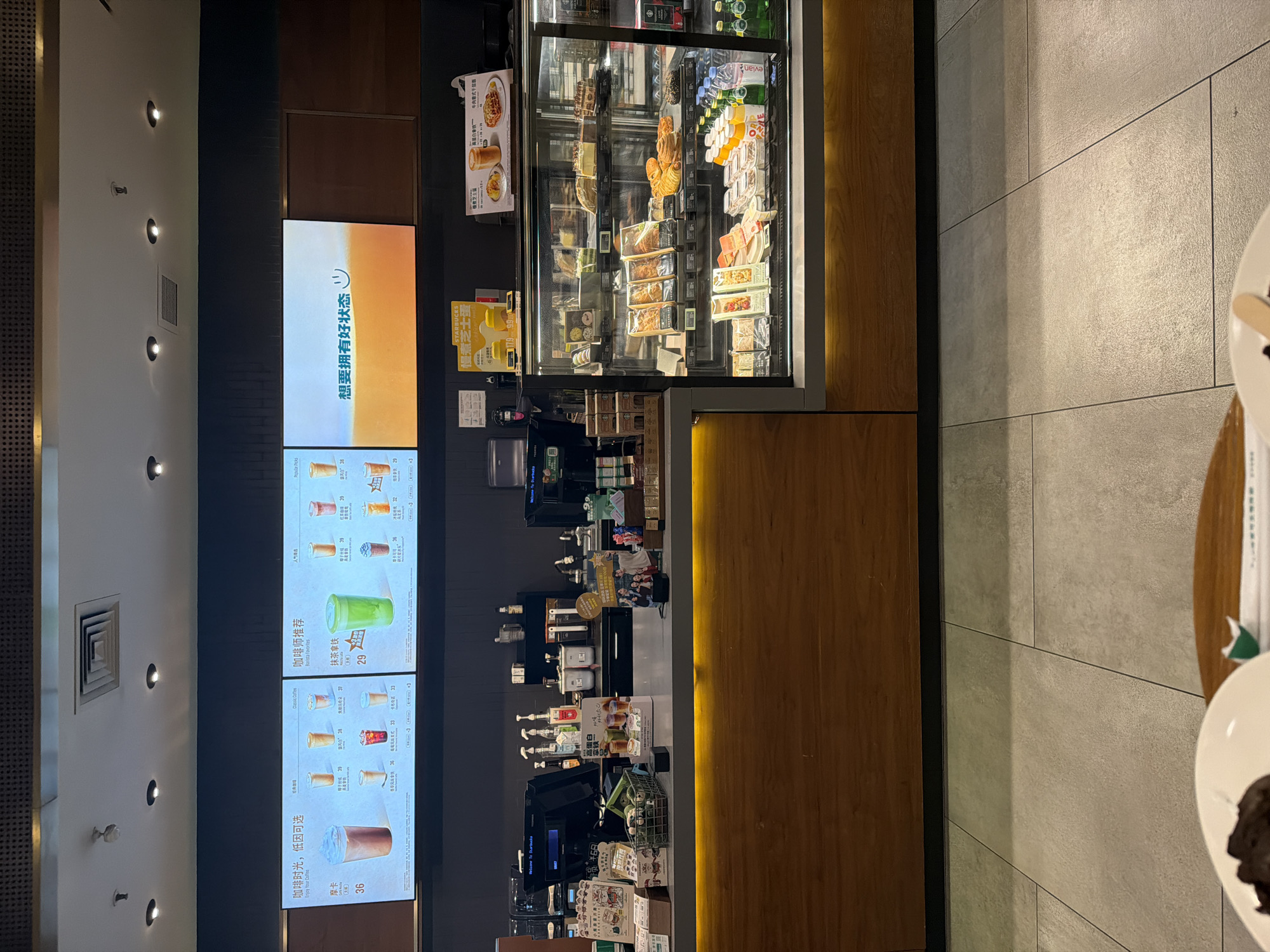 The image captures the interior of a brightly lit cafe, likely located in Yuhuatai District, China. The primary subject is a service counter with a prominent display case filled with various food and beverage items, including sandwiches, pastries, and bottled drinks like Evian water. Behind the counter, shelves hold additional products and equipment.
To the left of the counter, several vertical digital menu boards showcase a selection of drinks. One large screen displays Chinese text, "想要拥有好状态" (Want to have a good state/condition), accompanied by a smiling emoji. Below it, other screens list coffee and tea options with prices. Visible text includes "咖啡时光, 低因可选" (Coffee time, decaf optional), "Enjoy Your Coffee," "咖啡师推荐" (Barista recommendations), "Barista favorites," and specific drinks like "摩卡" (Mocha - 36), "抹茶拿铁" (Matcha Latte - 29), "高蛋白拿铁" (High Protein Latte - 36), "红茶拿铁" (Black Tea Latte - 39), "美式咖啡" (Americano - 29), and "拿铁" (Latte - 33). The Starbucks logo is visible on some promotional materials near the display case, along with items like "慢煮芝士蛋" (Slow-cooked Cheese Egg) and "高蛋白拿铁牛肉意式千层" (High Protein Latte Beef Italian Lasagna).
The scene is illuminated by overhead lights and strip lighting along the counter, creating a warm ambiance. The ceiling features recessed spotlights and an air vent. The floor and a wall to the right are covered in large, light-grey tiles. No people are visible, suggesting the image might have been taken during a quiet moment or focused on the cafe's offerings. The overall impression is that of a modern, well-stocked cafe ready to serve customers.