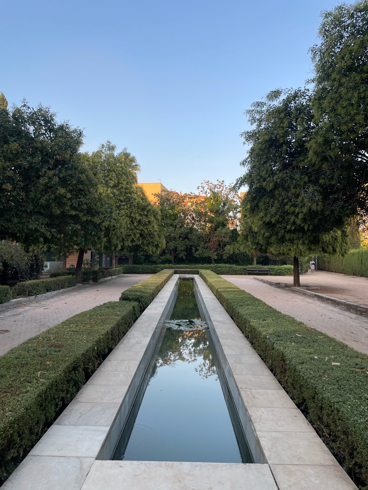 The image shows a long, rectangular reflecting pool in a public park. The pool is bordered by manicured green hedges and a paved walkway. Trees line the walkway, and in the background, buildings with orange roofs are visible. The sky is clear and blue. The scene appears to be from the Generalife gardens, part of the Alhambra complex in Granada, Spain. The time of day appears to be late afternoon or early evening, given the angle of the sunlight. One person, a woman in a patterned dress, is visible walking in the distance on the right side of the walkway.