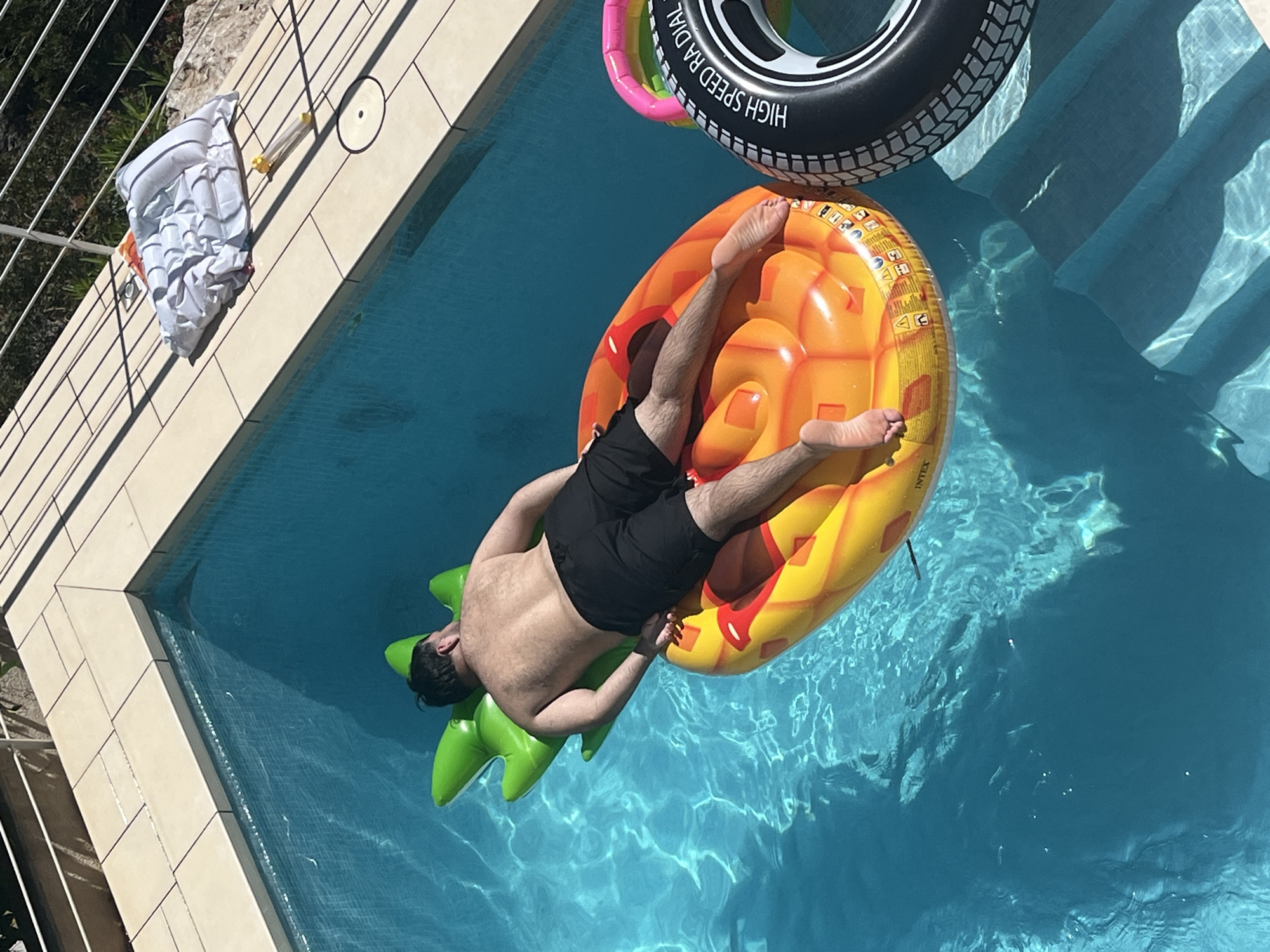 The scene depicts a man relaxing on a pineapple-shaped inflatable float in a swimming pool. He is wearing black swim trunks and is positioned face down on the float. Another inflatable, shaped like a tire, is also floating in the pool nearby. The pool is surrounded by tiled coping and a metal railing. A deflated inflatable is also on the pool's tiled coping, a small yellow air pump next to it. The location is likely a private residence in el Poble Nou de Benitatxell / Benitachell, Spain, based on the architectural style of the surrounding structures. The image is taken from an elevated perspective, looking down into the pool. The water is clear and reflects the sunlight.