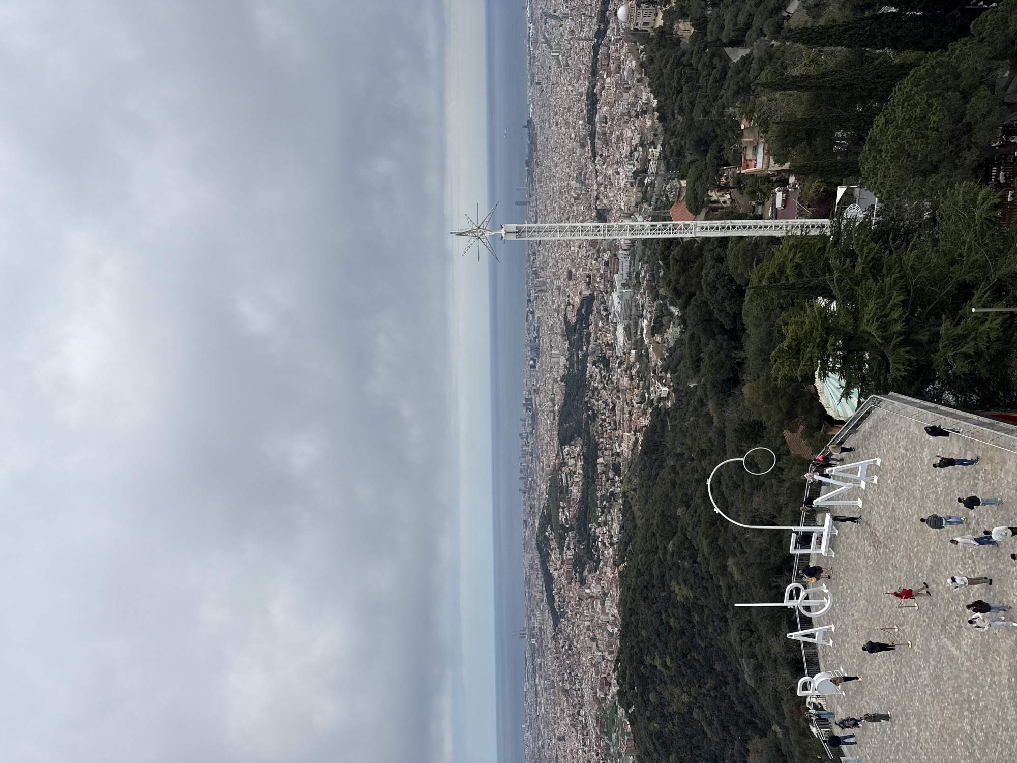 The image shows an elevated view of Barcelona, Spain, under an overcast sky. A tall, white metal structure resembling a tower extends from the foreground towards the horizon. The city stretches towards the Mediterranean Sea. On the right side of the frame, a viewing platform is visible with a large, white, cursive lettering that spells "BARCELONA," and people are standing and walking around on the platform. The terrain to the right includes dense green foliage, suggesting a hillside or park area.