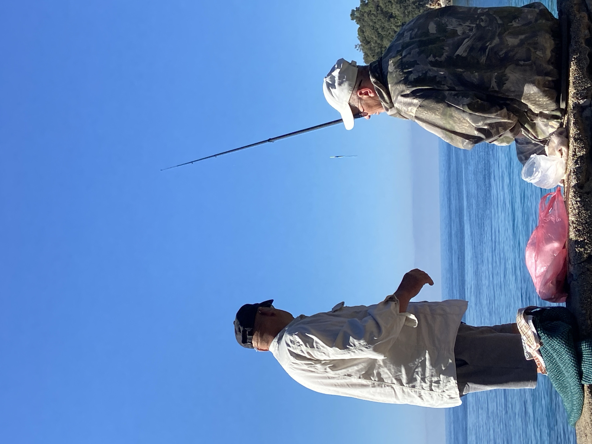 Two men are fishing. The man on the left is wearing a black baseball cap, a white shirt, and gray pants. The man on the right is wearing a white cap and a camouflage jacket. He is holding a fishing rod. There is a plastic bag next to him. The ocean is in the background. The location is Budva, Montenegro.