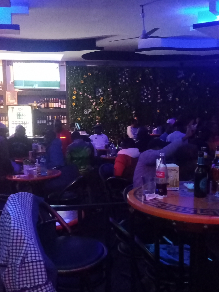 A dimly lit bar features a television above a stocked bar, a decorated wall, and a ceiling fan. Patrons are seated at tables, with drinks visible. The foreground shows empty tables and chairs. The scene is bathed in a mix of artificial and neon lighting, contributing to the overall ambiance.