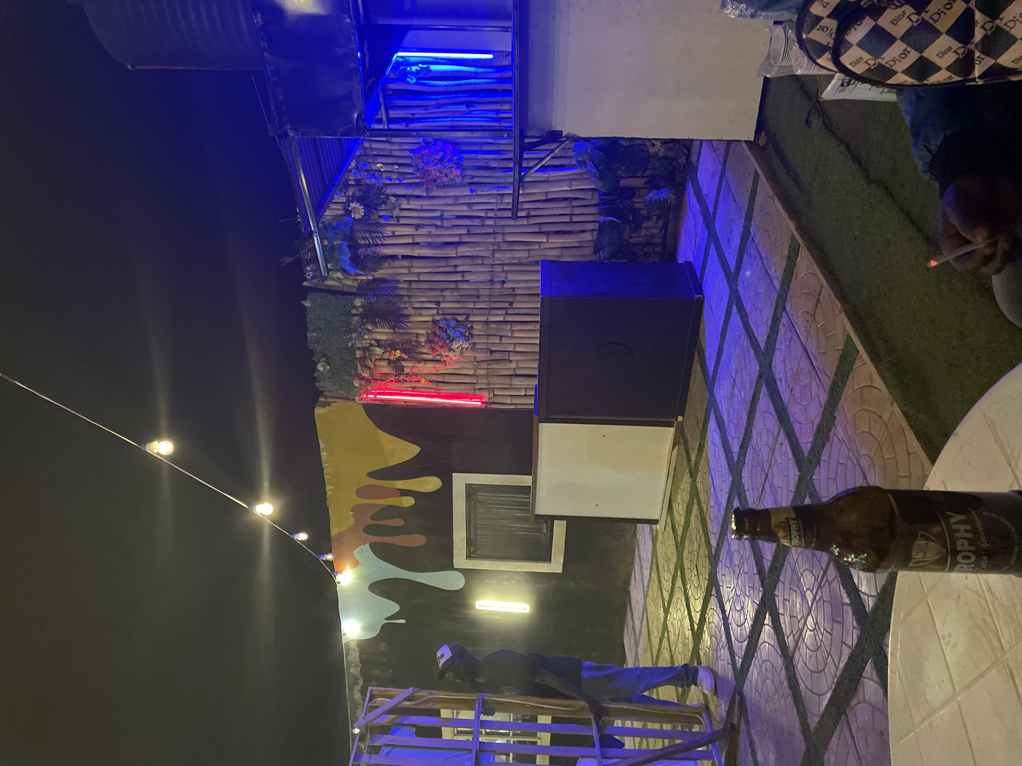 This image captures an outdoor evening or nighttime scene in Jos, Nigeria, depicting a casual social gathering spot. The setting is illuminated by various artificial lights, including string lights suspended against a dark sky, and vibrant blue and red neon lights.
The environment features a decorative wall constructed from bamboo-like panels, adorned with artificial greenery and floral arrangements. The ground is a combination of green artificial turf and geometrically patterned tiled paving, with blue light reflections on the tiles.
In the foreground on the right, a brown glass bottle of "TROPHY" beer is visible, resting on a white plastic table. Nearby, a person's hand is holding a lit cigarette. A bag with a distinctive black and white checkerboard pattern, featuring the word "Dior," is also visible, indicating a branded accessory.
Towards the bottom-left, a person wearing a dark top, blue trousers, and a cap is partially visible, standing behind a wooden crate structure. In the background, a wall is painted with an abstract mural featuring colorful, flowing shapes in yellow, brown, and light blue. A prominent black rectangular box, possibly a speaker or equipment housing, stands near the bamboo wall. The scene suggests a relaxed and social atmosphere at a bar, lounge, or outdoor event space.
