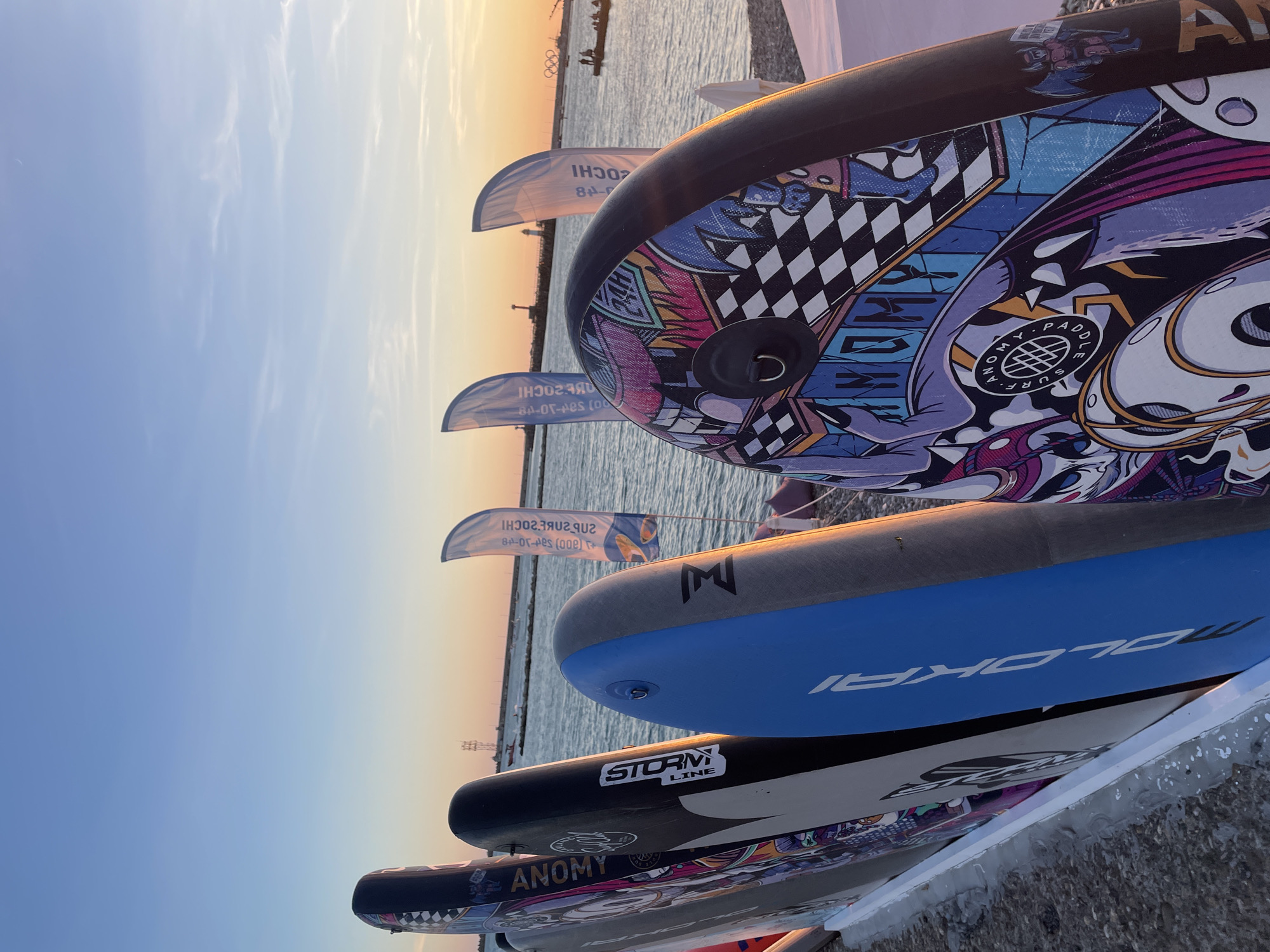 The image depicts a lineup of stand-up paddleboards on a gravelly shore with the Black Sea in the background. The sky is clear with a gradient from blue at the top to a warm orange and pink hue at the horizon, indicating either sunrise or sunset. Several flags, likely advertising, are planted in the sand or ground, some featuring Cyrillic text and graphics. In the distance, a breakwater or pier structure with a lighthouse is visible, as well as several small boats and the Olympic rings symbol, strongly suggesting the location is the city of Sochi, Russia, near the Olympic Park. The paddleboards are arranged vertically, leaning against a white support structure. They display various designs, including one with a black and white checkered pattern and cartoonish graphics, another with "ANOMY" branding, and a blue one with "MOLOKAI" visible. The overall scene conveys a sense of preparation for water sports or a display of equipment at a beachside venue.