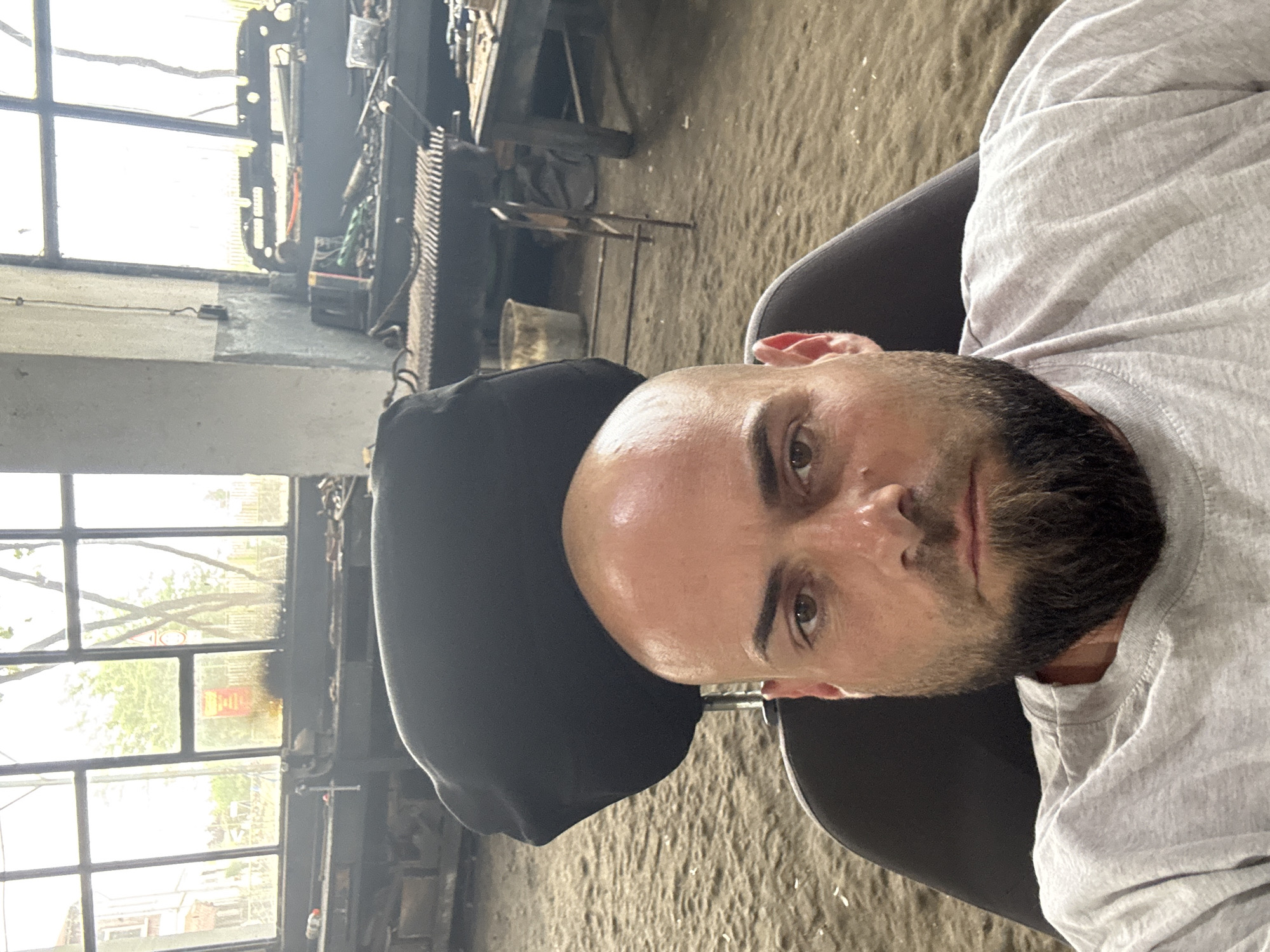 A medium shot captures a man in what appears to be an industrial workshop in Sarnevo, Bulgaria. The man, with a shaved head, a short dark beard, and dark eyes, is in the foreground, looking directly at the camera with a neutral expression. He is wearing a light gray t-shirt and is seated in a dark-colored chair, parts of which are visible behind his head and shoulders. There is a slight sheen on his forehead, possibly from light reflection or perspiration.
The background reveals a functional, cluttered workshop setting. Large, grimy windows with multiple panes dominate the left side of the frame, letting in bright daylight that suggests it is daytime. Faint greenery and distant structures are visible outside the windows. The rest of the workshop features workbenches or shelves laden with various tools, metal objects, and dark machinery. A large, dark piece of equipment with a serrated edge is noticeable on the right side of the background, and a bucket is on the floor. The floor itself is a rough, dusty concrete or dirt surface. The overall environment conveys a working industrial space, possibly for metalworking or manufacturing. There is no visible text in the image. The man's direct gaze suggests he is posing for the photograph.