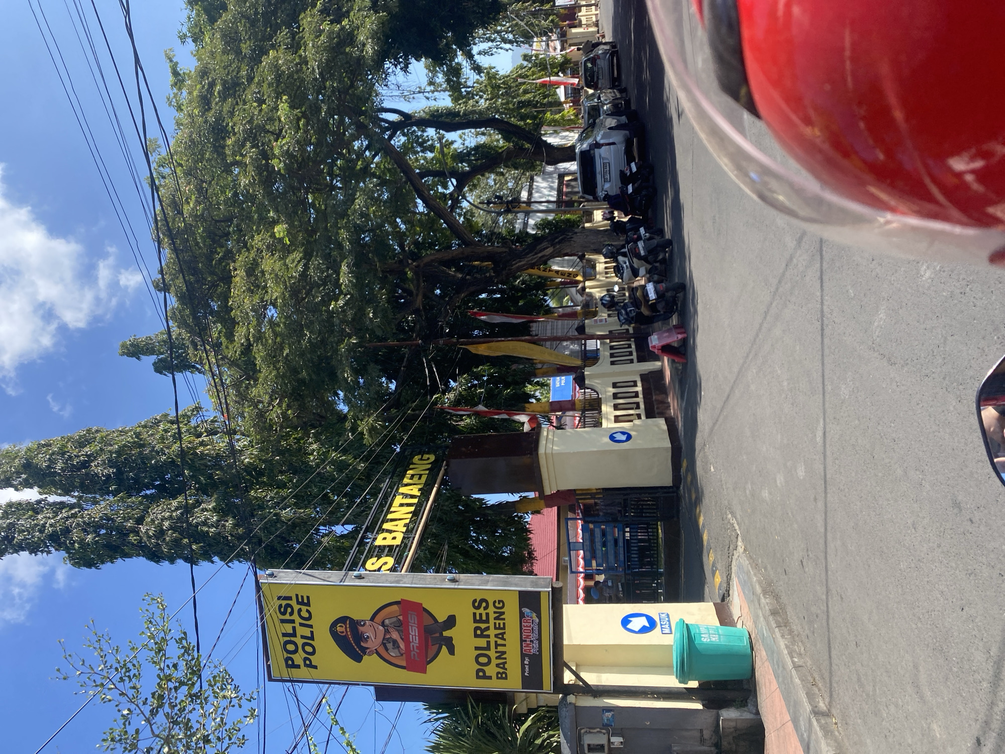 The image depicts the entrance to the Polres Bantaeng, which is the police station in Bantaeng, Indonesia. A prominent yellow sign with "POLISI POLICE" and a cartoon police officer is visible. Below this, it reads "POLRES BANTAENG". The entrance gate is partially visible with blue bars. Several motorcycles are parked on the side of the road in front of the building, and a white SUV is also parked nearby. The scene appears to be a sunny day with a clear blue sky and some clouds. Tall trees line the street, providing shade. A green trash bin with "SAMPA KEPI" written on it is placed on the sidewalk to the left of the entrance. Yellow and white striped speed bumps are present on the road leading to the gate. The perspective suggests the photo was taken from a vehicle, possibly a motorcycle, as a red helmet is visible in the foreground.